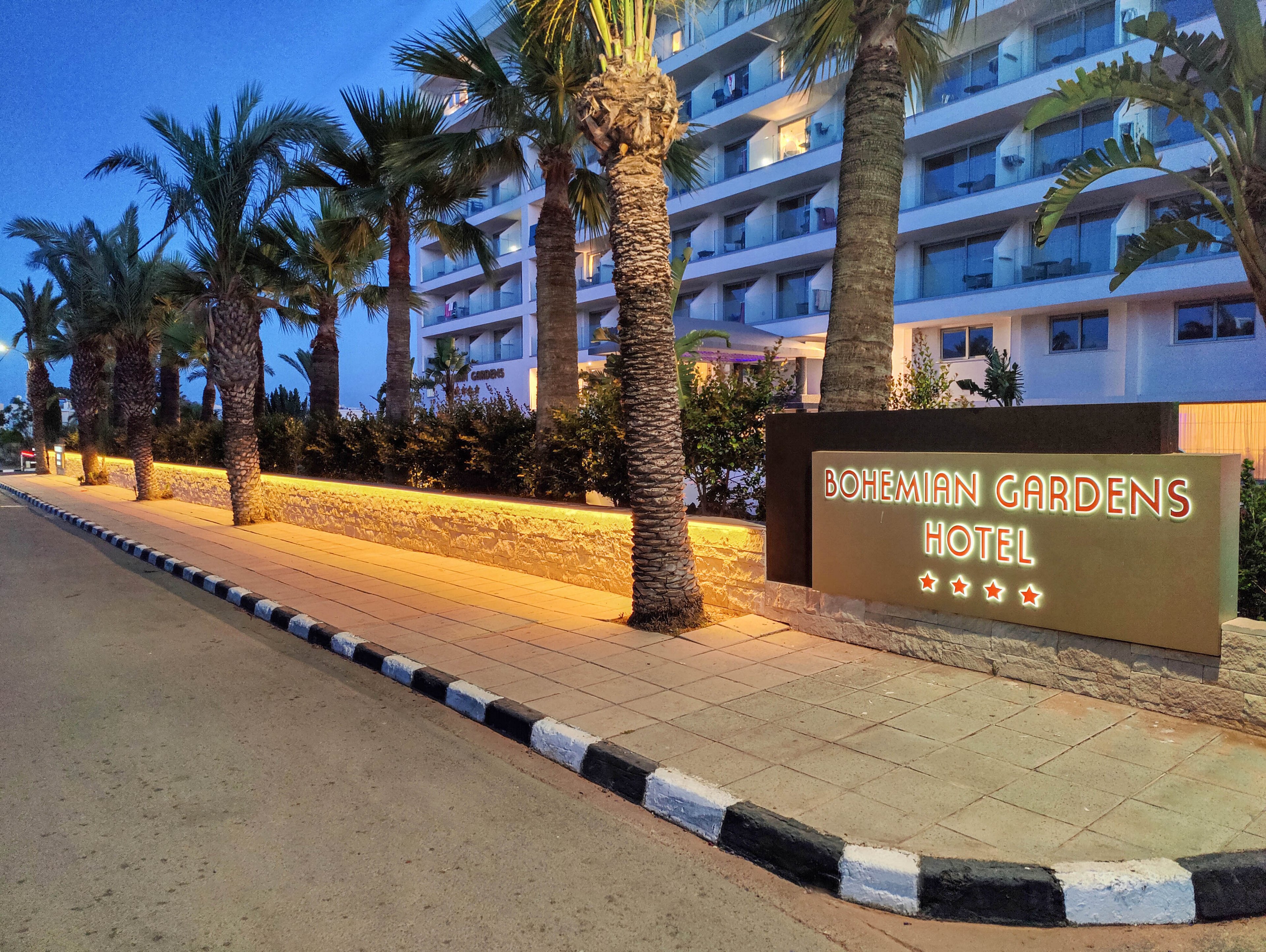 A modern hotel entrance is shown at dusk with a lit sign reading "Bohemian Gardens Hotel". Palm trees line the sidewalk beside the building, creating a resort-like atmosphere. Warm lighting along the low stone wall contrasts with the cool blue evening sky. The scene suggests a welcoming, tranquil vacation destination.