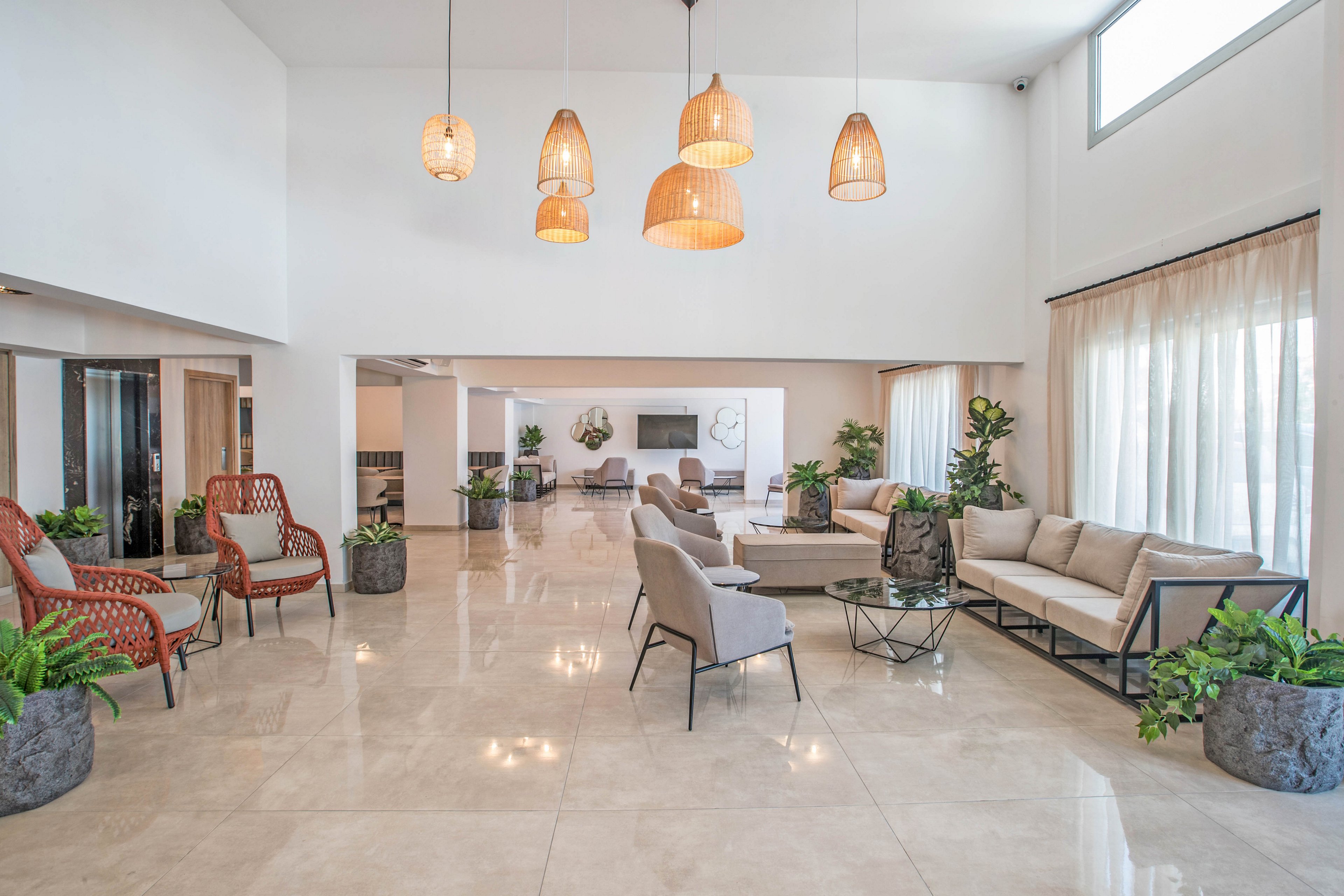 Spacious contemporary lobby interior featuring multiple seating areas with sofas and armchairs. Large floor-to-ceiling windows with sheer curtains allow abundant natural light into the room. Pendant lights with warm tones hang from the high ceiling, complementing the neutral tile flooring. Green potted plants and minimalist decor add a fresh, welcoming atmosphere to the modern space.