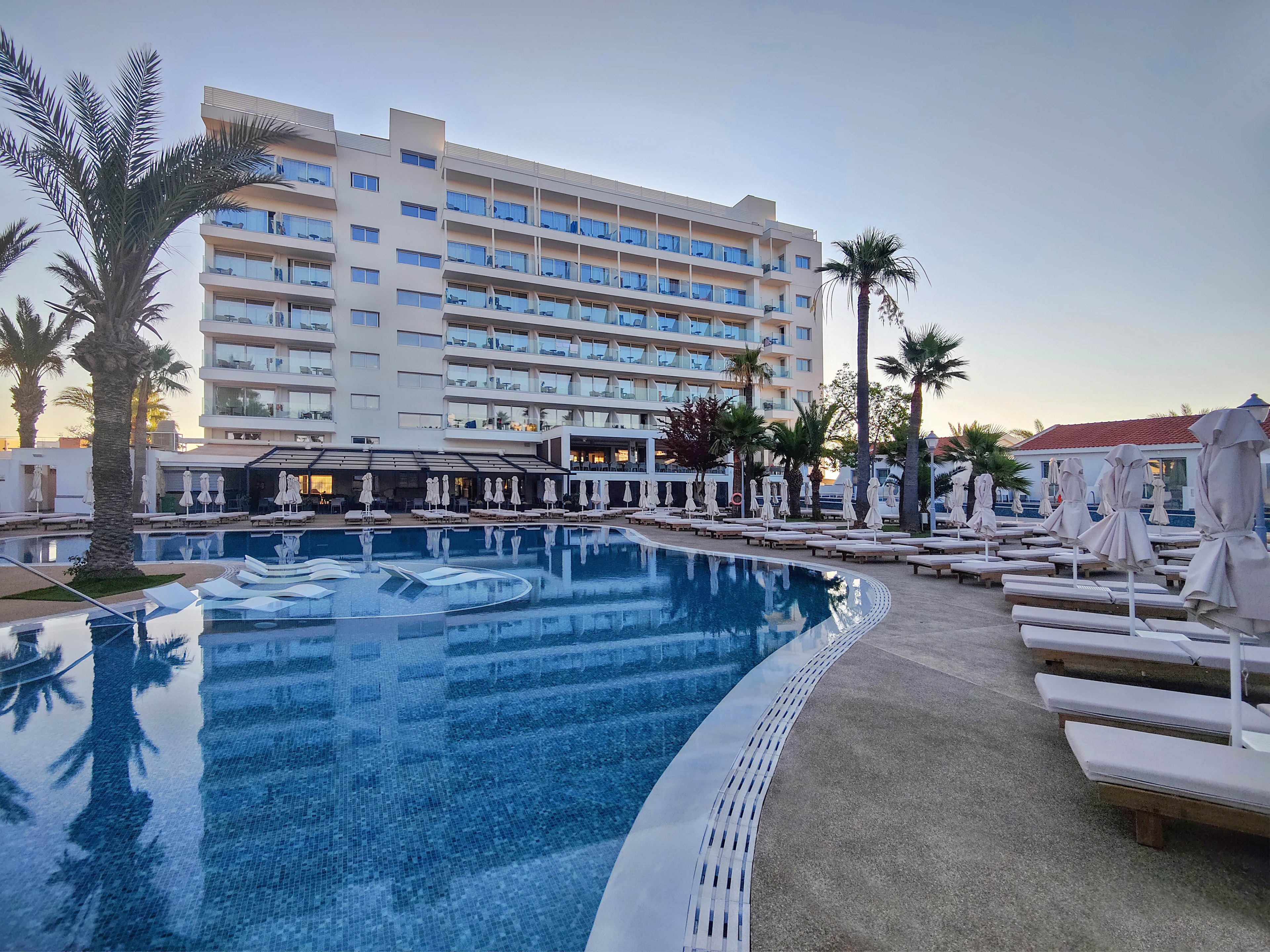 A large modern hotel overlooks a curved outdoor swimming pool at dusk. Numerous white sun loungers and closed umbrellas line the poolside, creating a calm and orderly scene. Tall palm trees and landscaped greenery add a tropical resort atmosphere. The sky is softly lit by the setting sun, reflecting in the still blue water and glass balconies of the building.