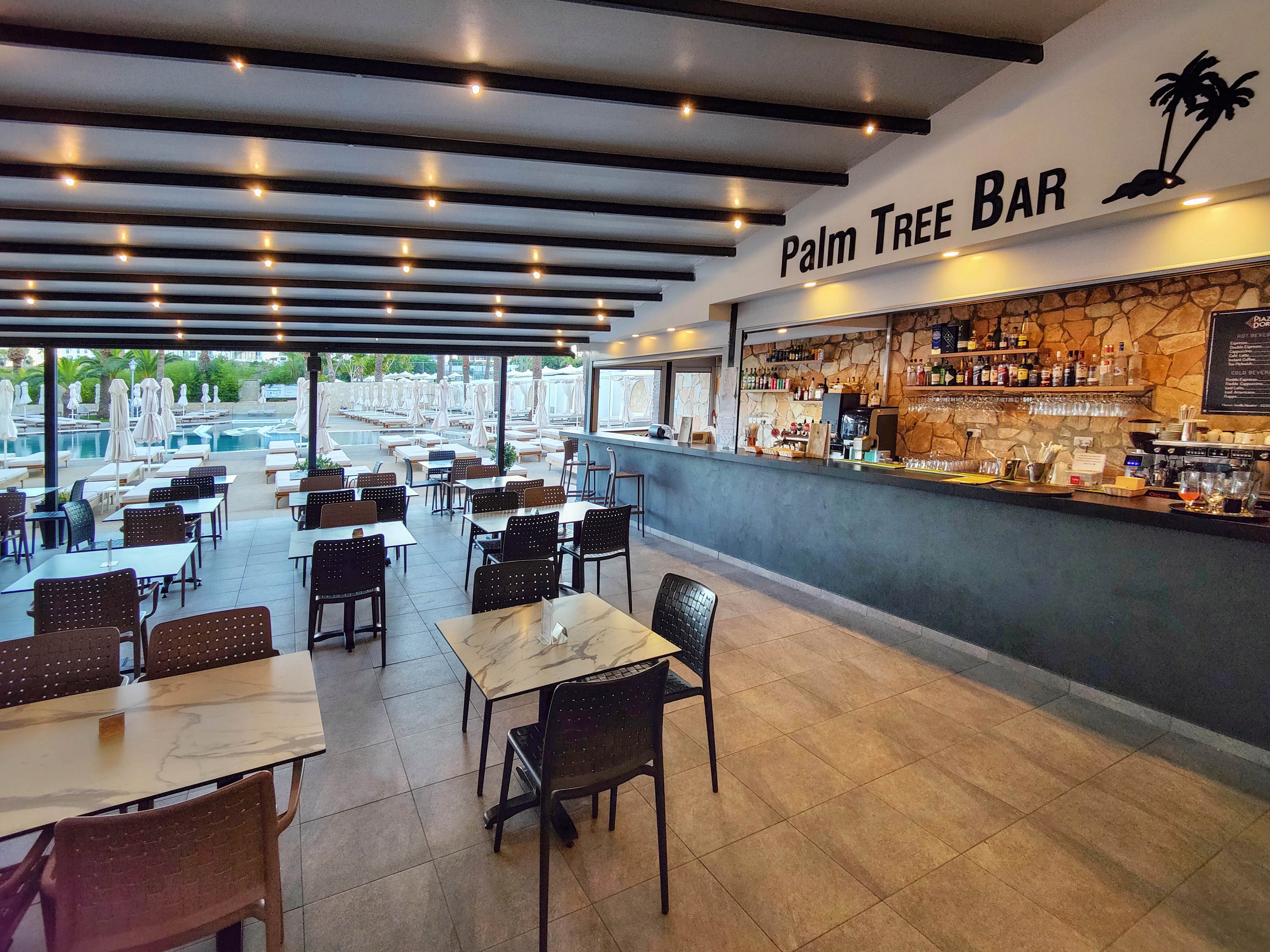 Spacious bar interior with the name Palm Tree Bar displayed above a long counter. The setting overlooks an outdoor pool area with sun loungers and umbrellas visible through large open windows. Neatly arranged tables and chairs create an inviting, casual dining and drinking space. Warm ceiling lights and stone wall accents contribute to a relaxed, holiday atmosphere.