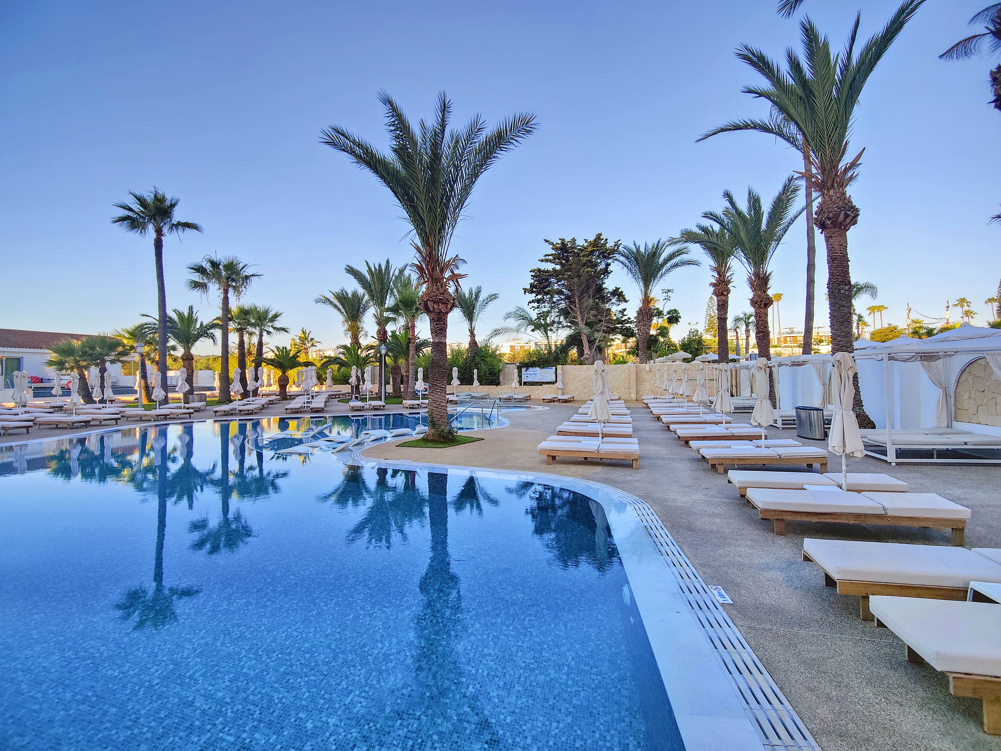 A large, tranquil swimming pool is surrounded by rows of white sun loungers and cabanas at a luxury resort. Tall palm trees line the poolside, reflecting clearly in the calm blue water. The setting appears quiet and empty, suggesting early morning or off-peak relaxation time. The overall scene conveys a serene, upscale vacation atmosphere under a clear sky.