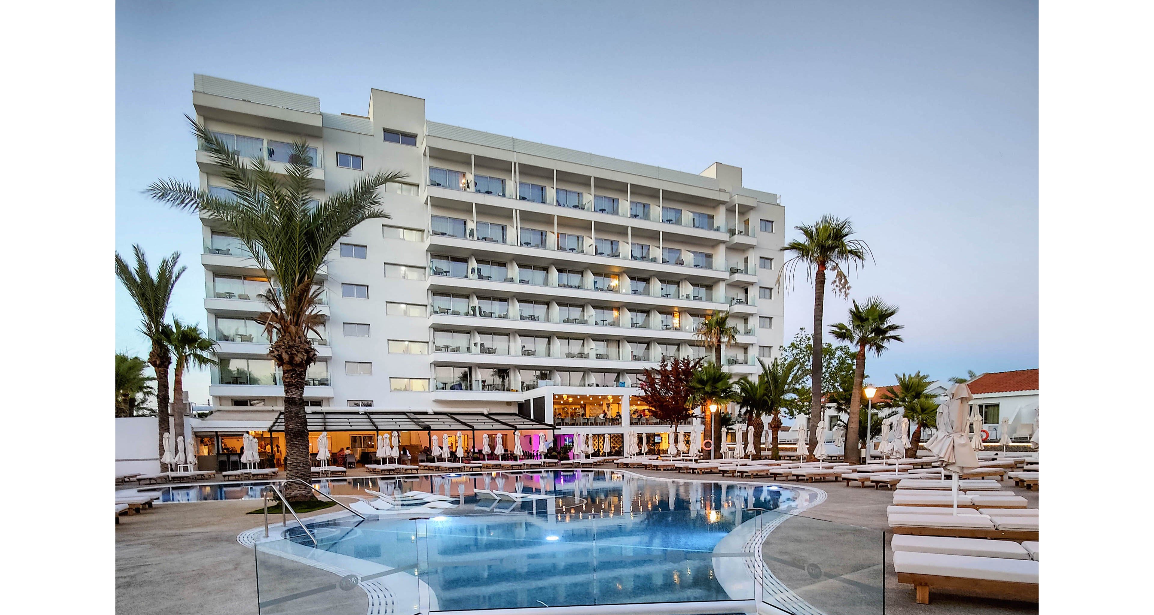 A contemporary multi-story hotel overlooks a curved outdoor swimming pool surrounded by sun loungers. Palm trees and landscaped greenery frame the pool deck, creating a relaxed resort atmosphere. The building’s balconies face the pool area, suggesting rooms with views of the courtyard and water. Soft evening light and a clear sky enhance the tranquil vacation setting.