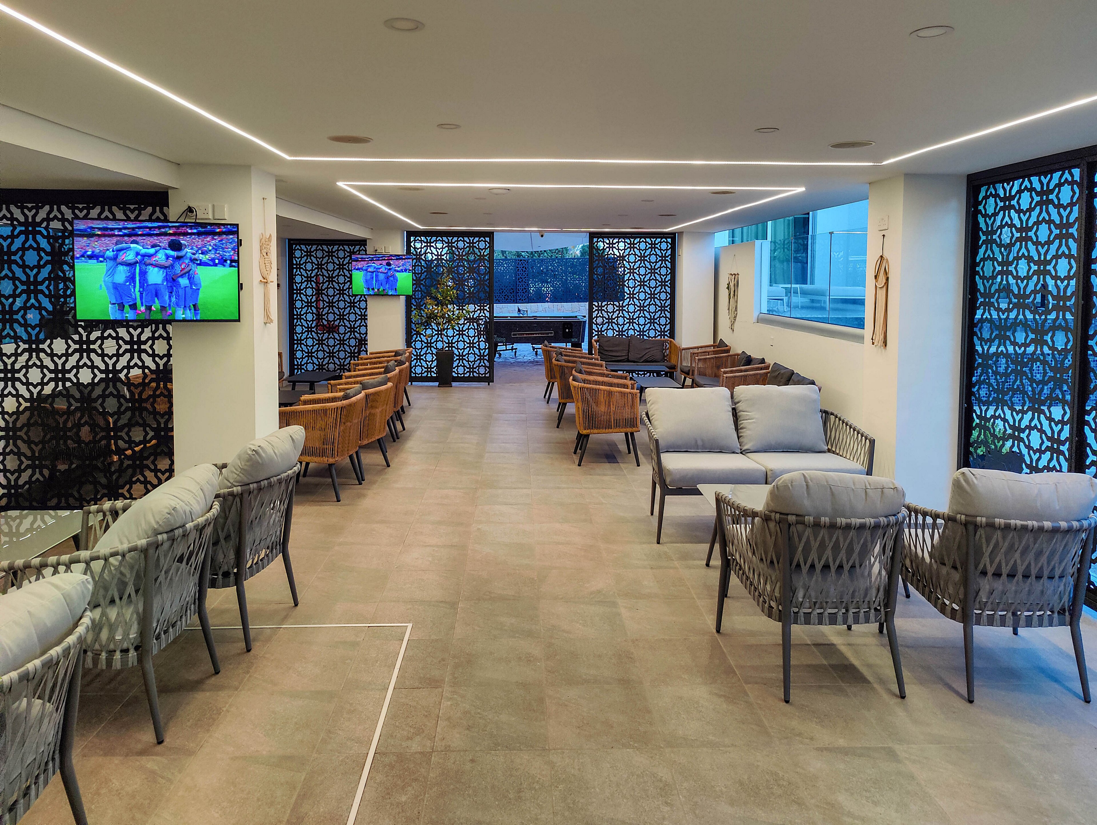 A contemporary indoor lounge features cushioned chairs and sofas arranged in neat rows. Decorative lattice screens line the walls, adding texture and privacy to the bright space. Two wall-mounted televisions display a sports match, providing entertainment for guests. Large windows at the far end let in natural light and offer a view outside, enhancing the relaxed, upscale atmosphere.