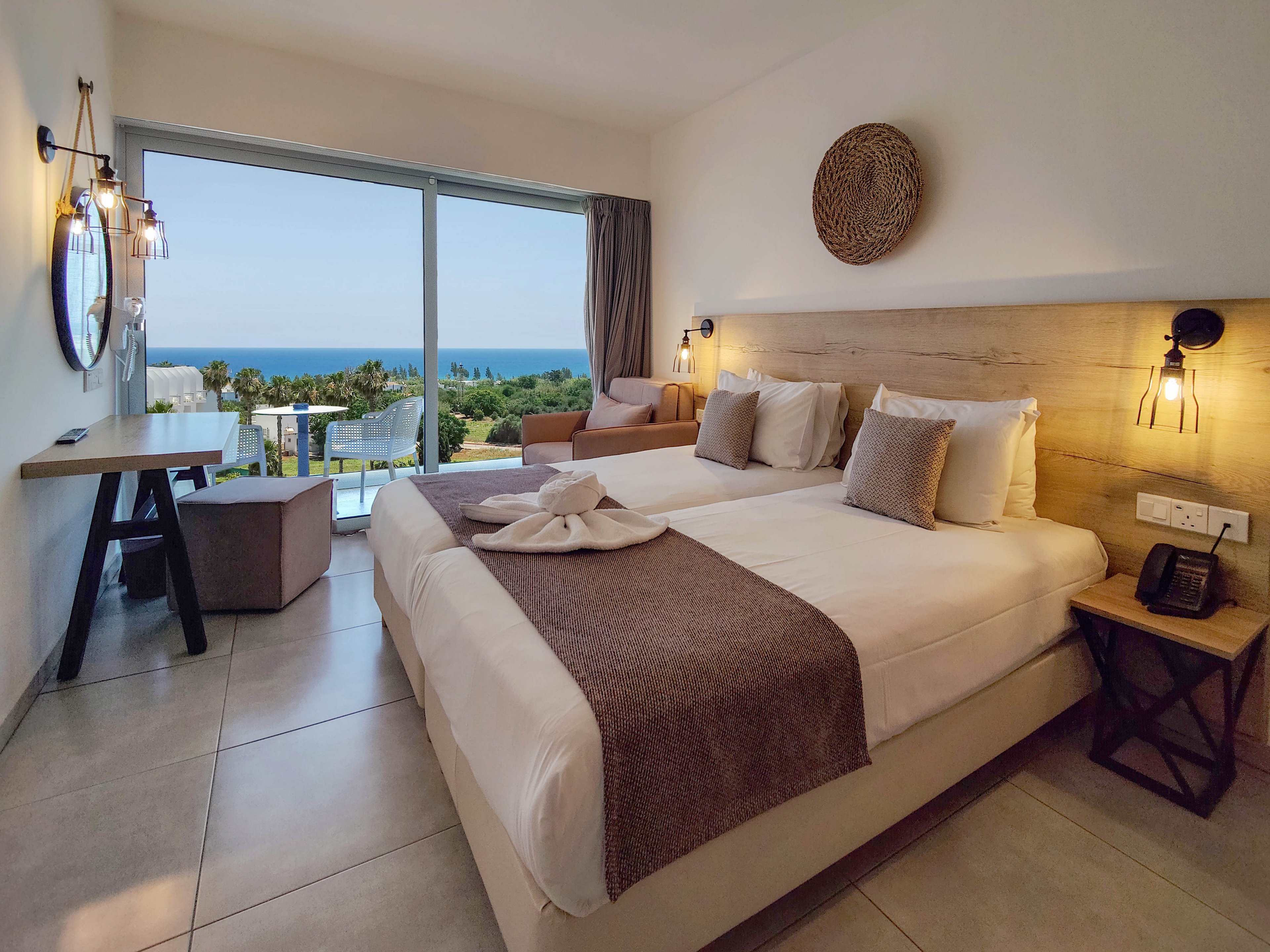 Contemporary hotel room features two neatly made twin beds with neutral linens and soft cushions. Large sliding glass doors open to a balcony overlooking lush greenery and a distant ocean horizon. Warm wall sconces, a woven wall decoration, and minimalist furniture create a cozy, stylish atmosphere. The space feels bright and inviting, ideal for a relaxing seaside vacation stay.