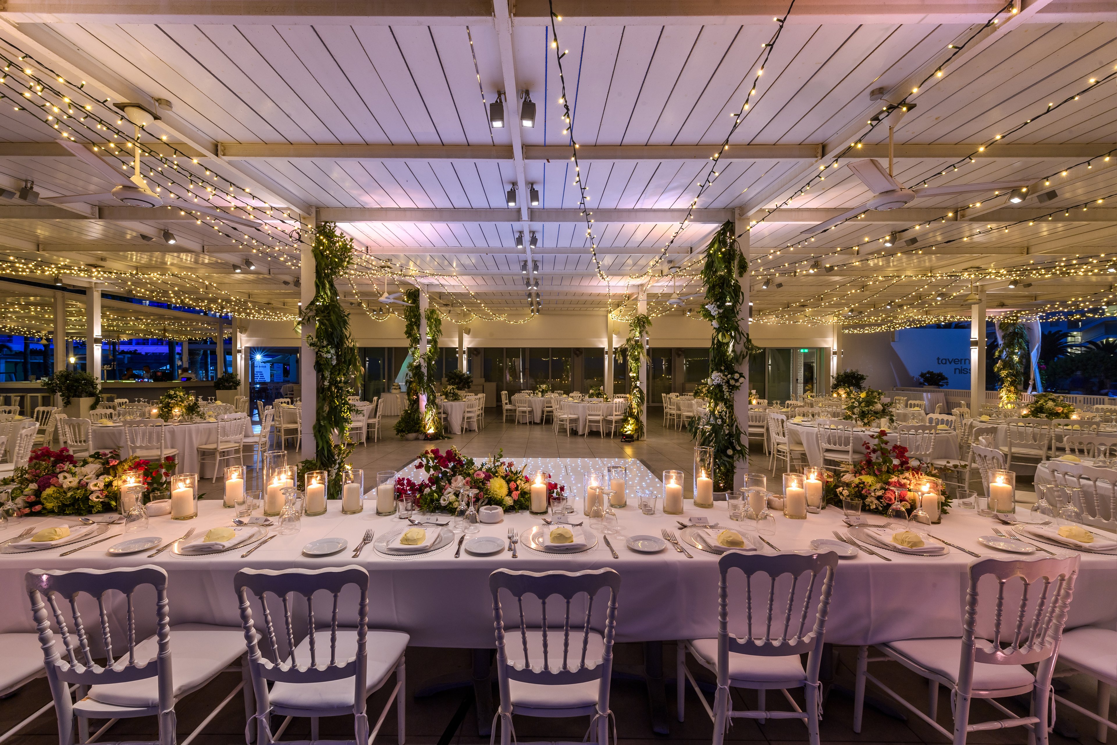 A long banquet table is set for an elegant event, decorated with floral centerpieces and glowing candles. White chairs and neatly arranged place settings create a formal wedding or celebration atmosphere. Overhead, string lights and greenery-wrapped columns add warmth and charm to the open-air venue. The scene is captured at dusk, highlighting the romantic lighting and spacious layout.