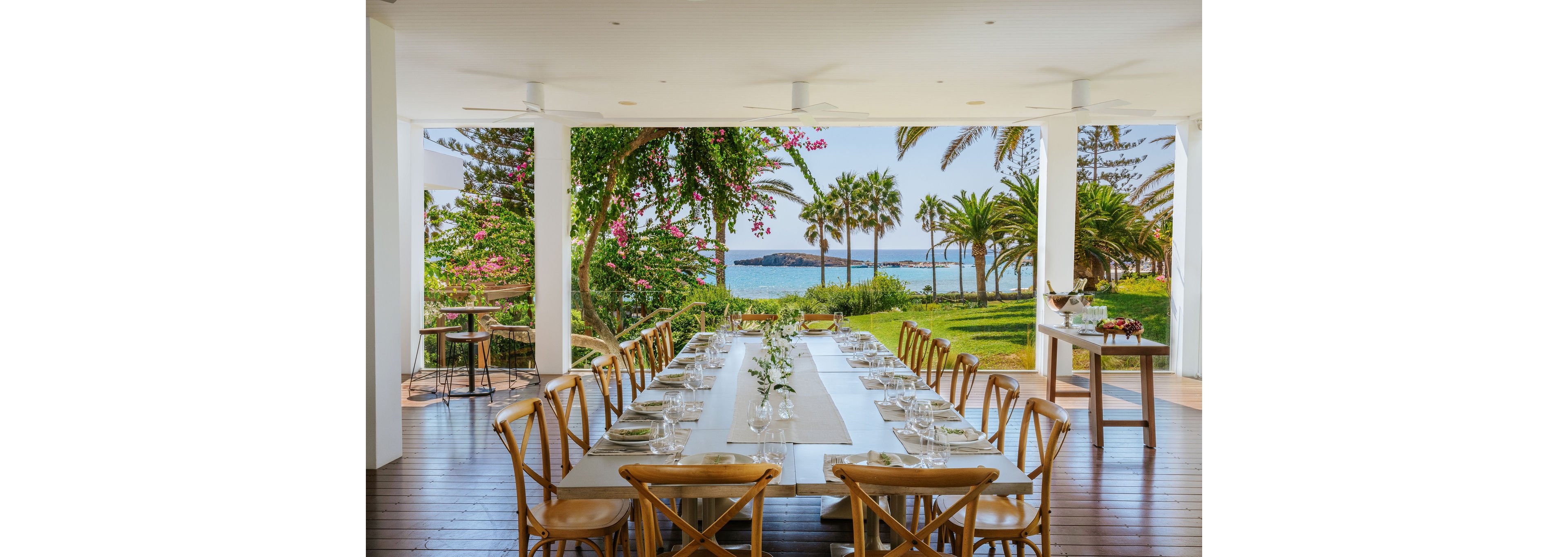 A long, elegantly set dining table stands on a covered terrace overlooking a lush garden and the ocean. Wooden chairs line both sides of the table, which is arranged with plates, glasses, and simple floral centerpieces. Bright greenery, palm trees, and blooming flowers frame the open view toward the blue water and distant shoreline. The scene feels airy and inviting, ideal for a coastal gathering or celebration.