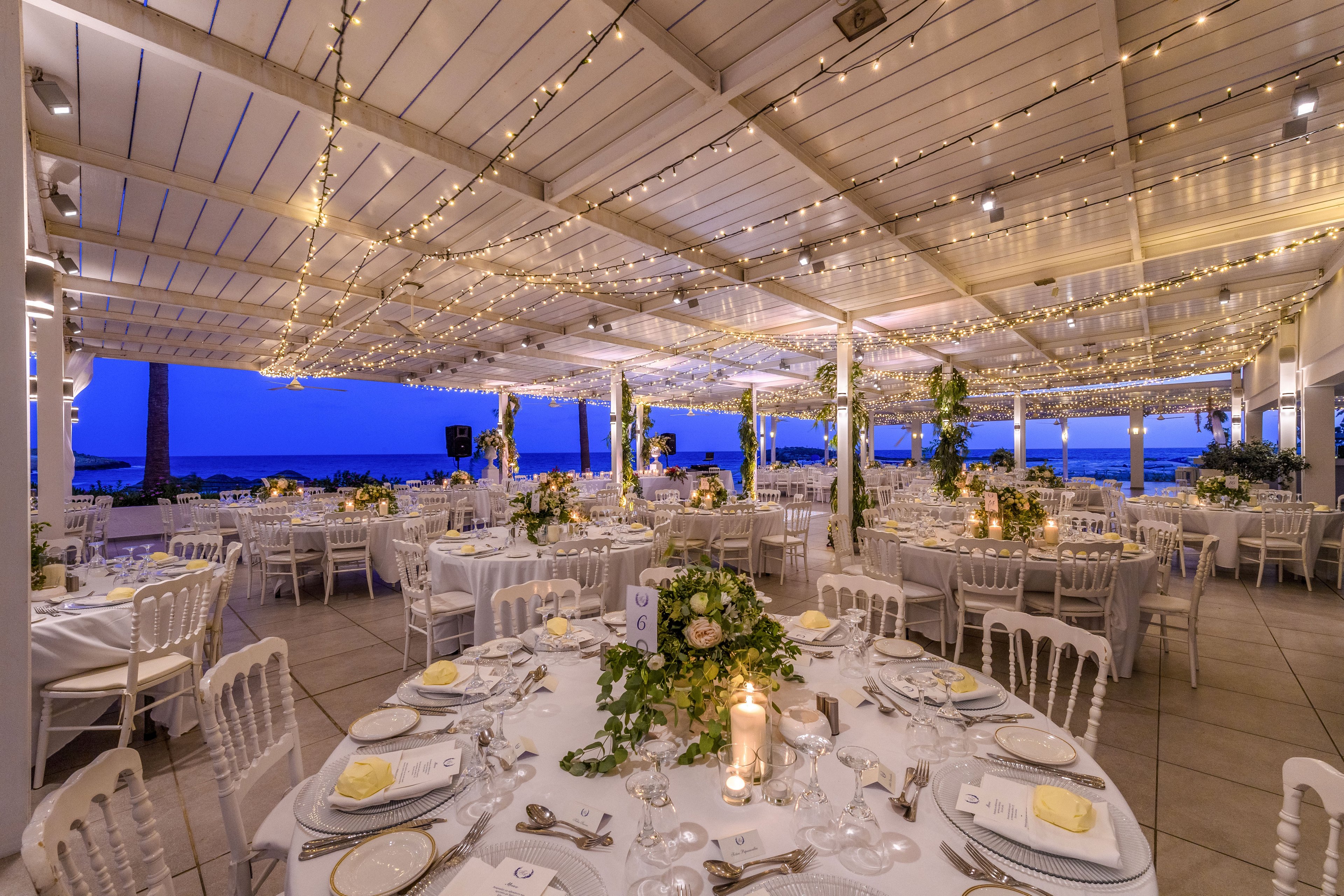An elegant wedding reception is set under a covered terrace overlooking the sea at dusk. Round tables are arranged with white linens, fine tableware, and floral centerpieces, creating a sophisticated atmosphere. Warm string lights and candles illuminate the space, contrasting with the deep blue evening sky. The scene feels romantic and festive, ready to welcome guests for a formal celebration.