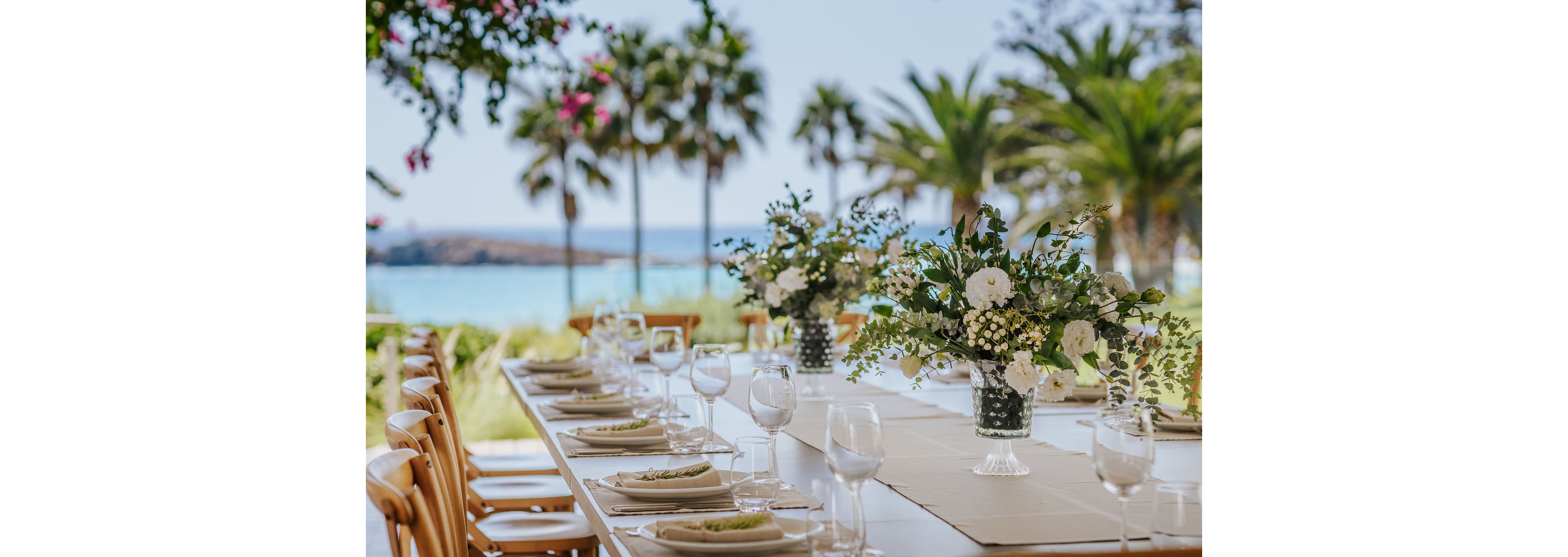 A long dining table is beautifully arranged outdoors with white plates, glassware, and neatly folded napkins. Lush floral centerpieces featuring white flowers and greenery run along the middle of the table. In the background, palm trees and a bright blue sea create a tropical coastal atmosphere. The scene suggests a sophisticated celebration or event in a sunny, relaxed setting.