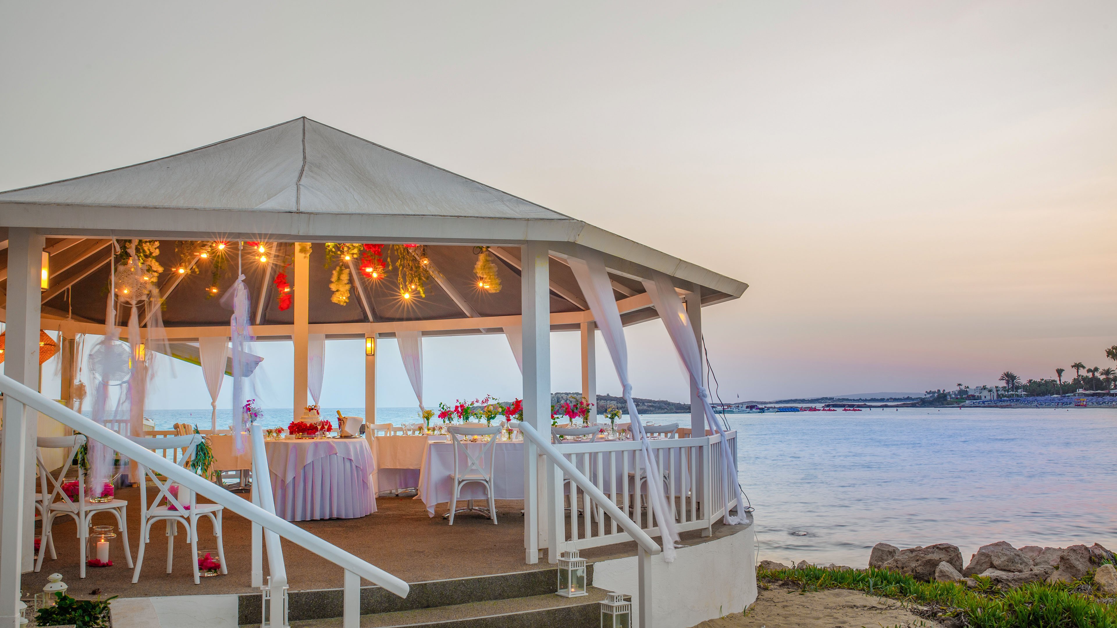 A white gazebo is beautifully decorated for an event beside a calm seaside at sunset. Tables inside the gazebo are arranged with flowers, candles, and colorful decorations, suggesting a wedding or special celebration. Soft string lights hang from the ceiling, creating a warm and romantic atmosphere. The tranquil water and pastel sky provide a serene coastal backdrop to the festive scene.