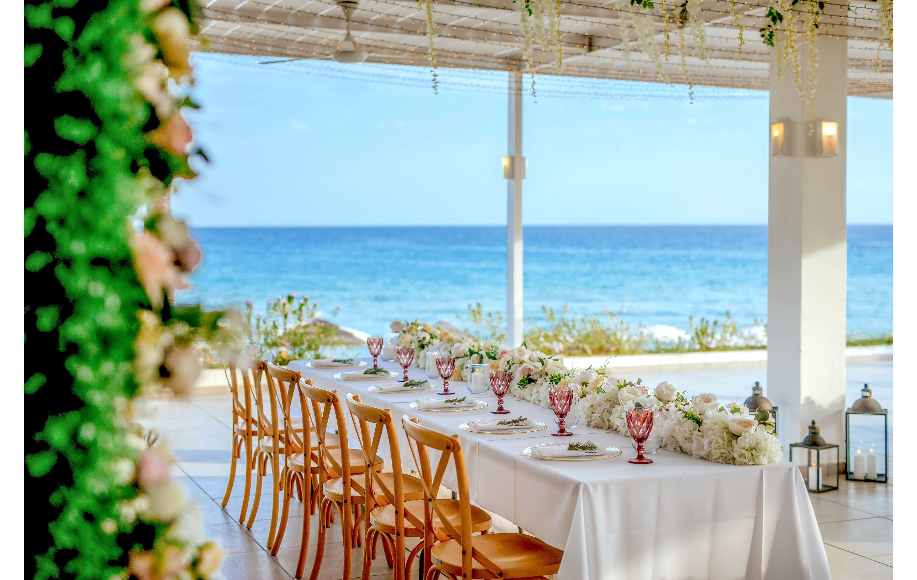 A long white-clothed table is set for a formal event beside a bright blue ocean. Wooden chairs line both sides of the table, which is decorated with white floral arrangements and pink glass goblets. The open-air venue features a shaded pergola ceiling, hanging greenery, and soft wall lights. Lanterns and coastal scenery in the background create a romantic, tranquil atmosphere.