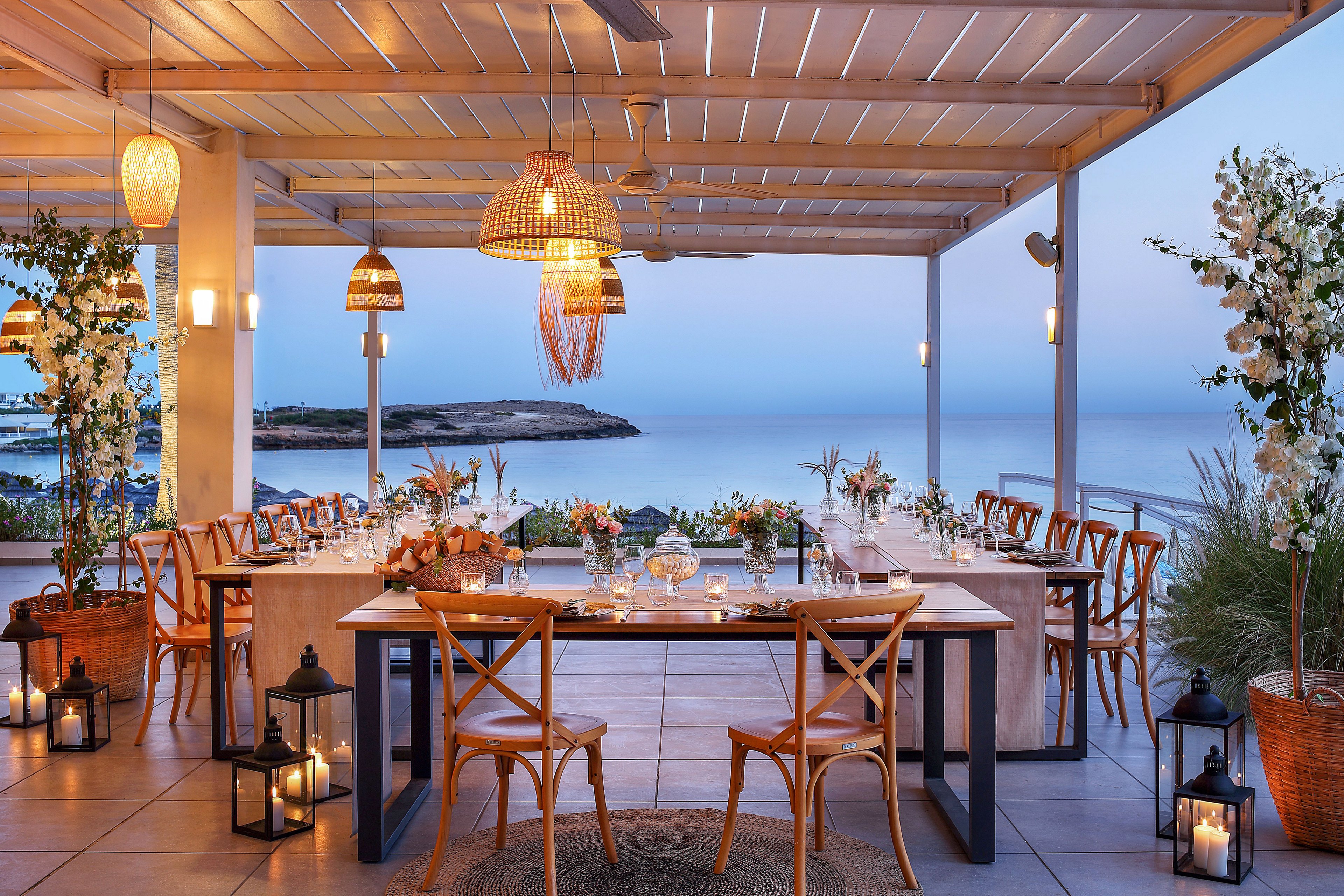 A long wooden table is beautifully set for dinner on an open-air terrace overlooking a calm sea at dusk. Wooden chairs, glassware, and floral centerpieces create a refined yet relaxed atmosphere. Warm lanterns, candles, and woven pendant lights add soft, ambient lighting to the scene. Potted plants and coastal views frame the space, suggesting a romantic or celebratory gathering by the water.
