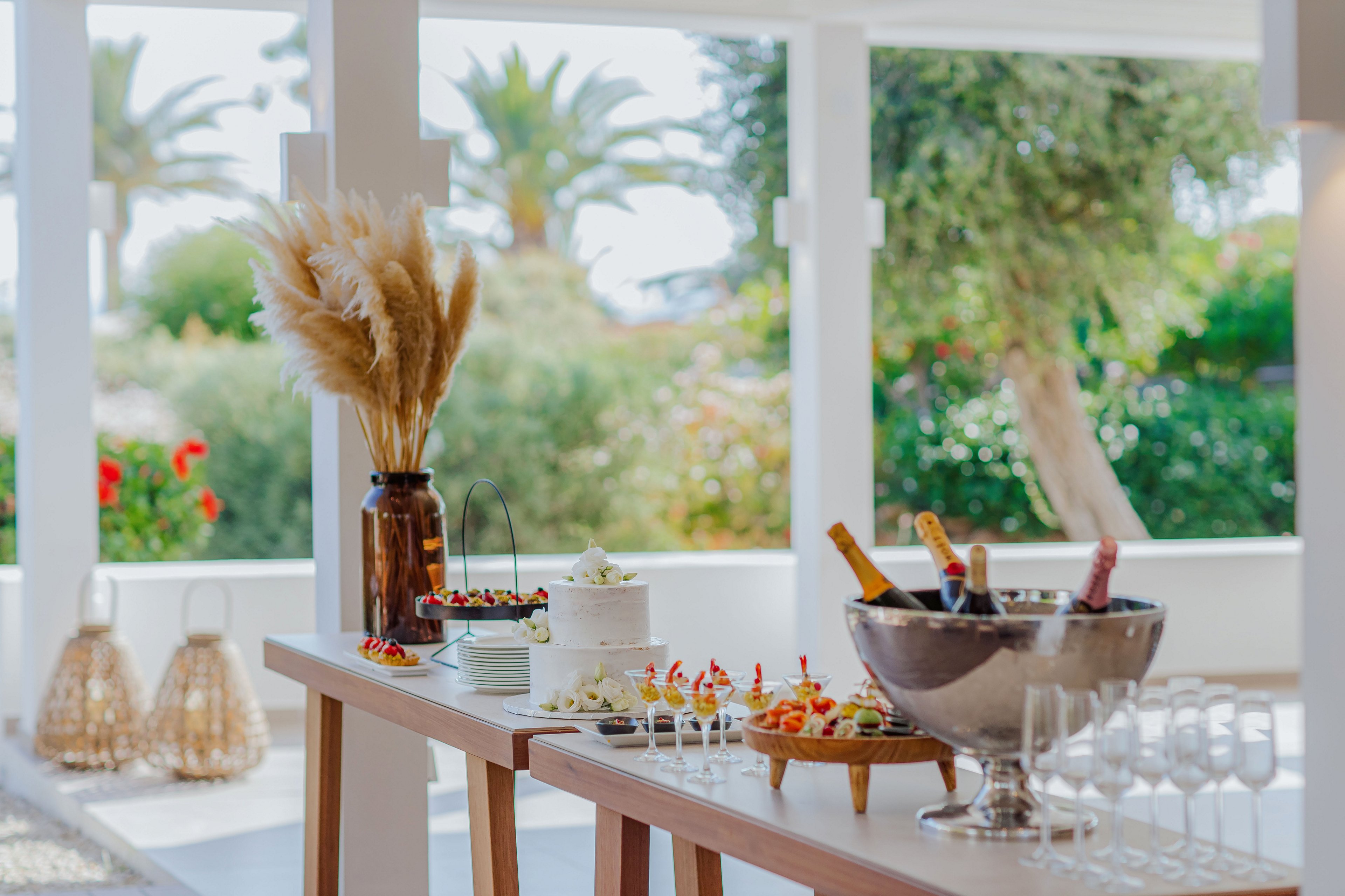 An elegant buffet table is set up outdoors with a variety of appetizers and a white frosted cake as the centerpiece. A large metal ice bucket holds several bottles of chilled champagne beside rows of empty wine glasses. Decorative pampas grass in a brown vase and woven lanterns enhance the stylish presentation. The bright terrace setting, greenery, and palm trees in the background create a relaxed, summery atmosphere.