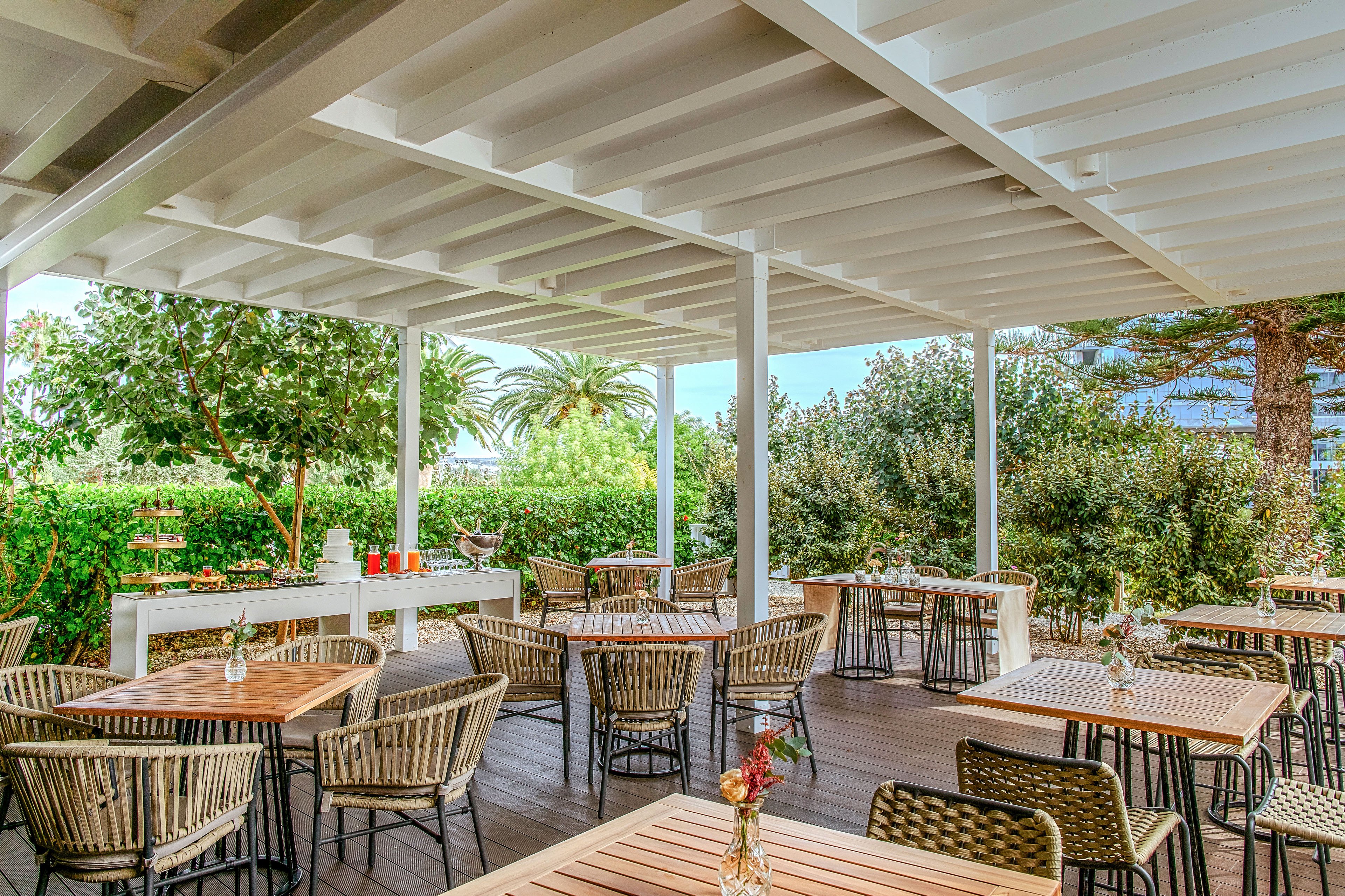 An open-air café patio features neatly arranged wooden tables and woven chairs under a white pergola roof. Small vases with flowers decorate each table, adding a touch of color and charm. The setting is surrounded by lush greenery, including trees and hedges, with a glimpse of palm trees in the background. Bright natural light and clean lines create a relaxed, inviting atmosphere suitable for dining or social gatherings.