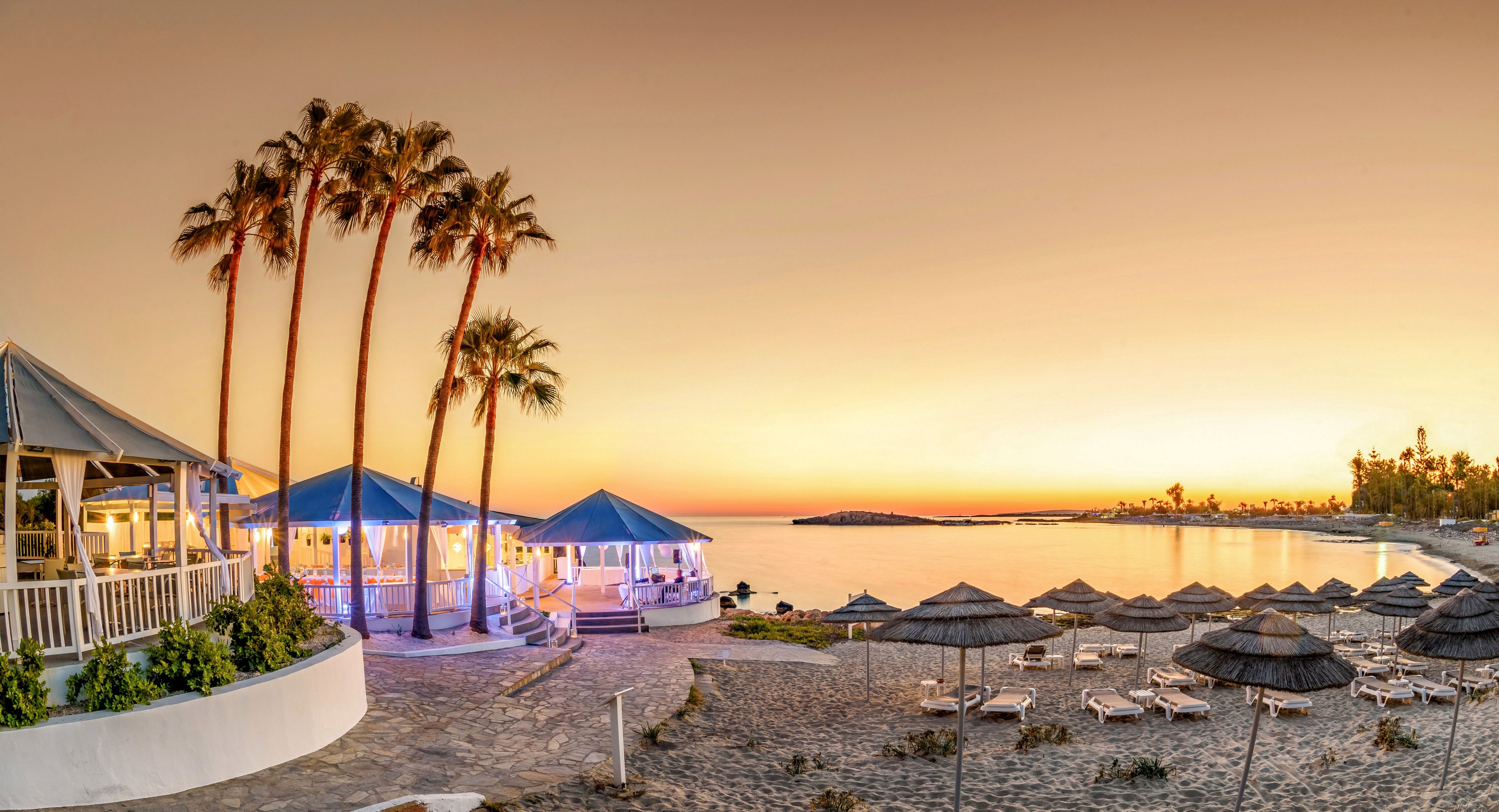 A tranquil seaside resort is captured at sunset, with tall palm trees framing open-air pavilions and beach bars. The sky glows in warm golden tones, reflecting softly on the calm sea. Straw umbrellas and sun loungers line the sandy beach, suggesting a relaxed vacation atmosphere. Soft artificial lighting from the buildings contrasts with the natural twilight, enhancing the serene coastal scene.