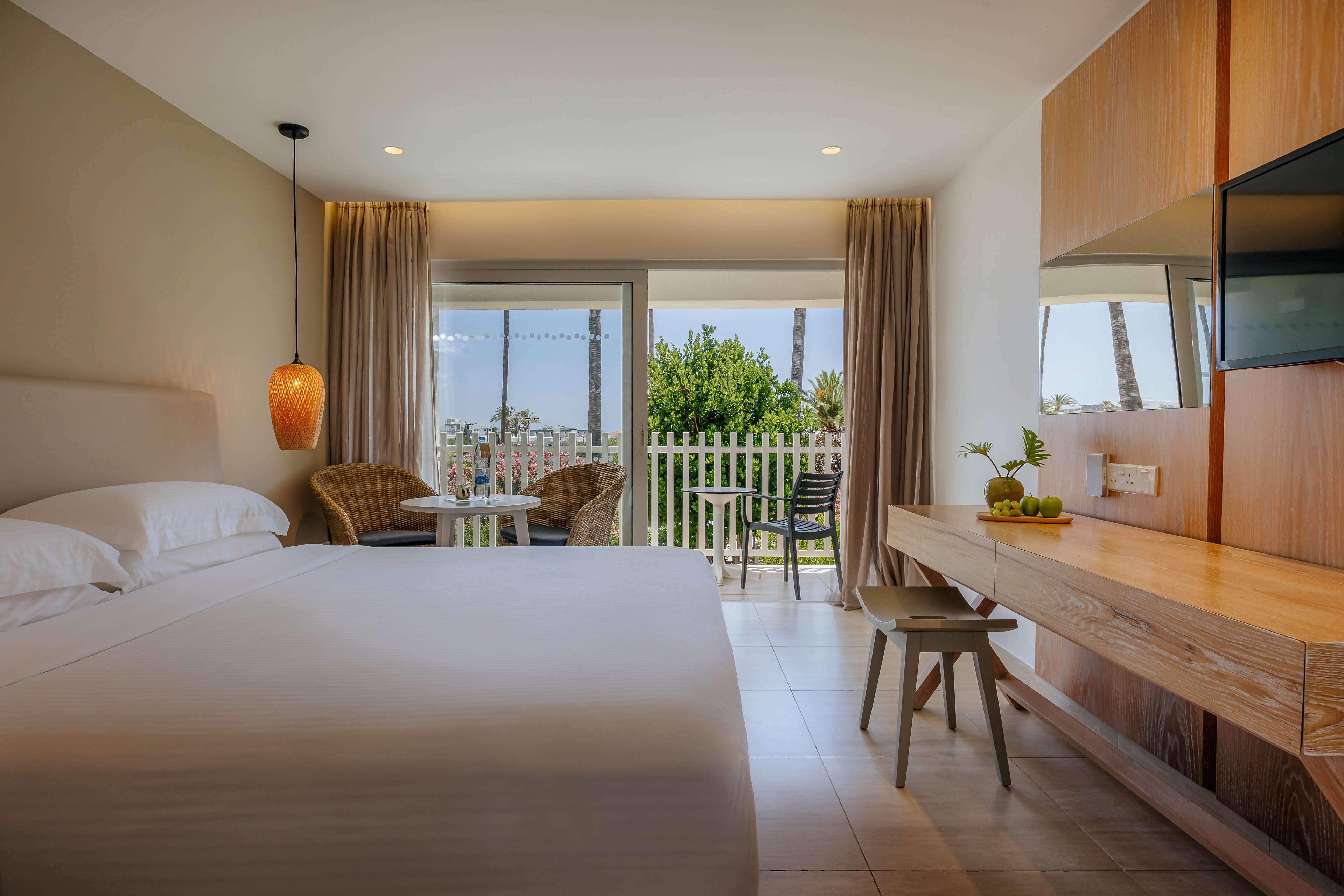 Contemporary hotel bedroom featuring a large neatly made bed and minimalist wooden furnishings. A sliding glass door opens to a balcony with outdoor chairs and a table overlooking greenery and palm trees. Warm lighting from a hanging lamp and neutral curtains create a cozy, inviting atmosphere. A wall-mounted television and small desk area complete the comfortable vacation setting.