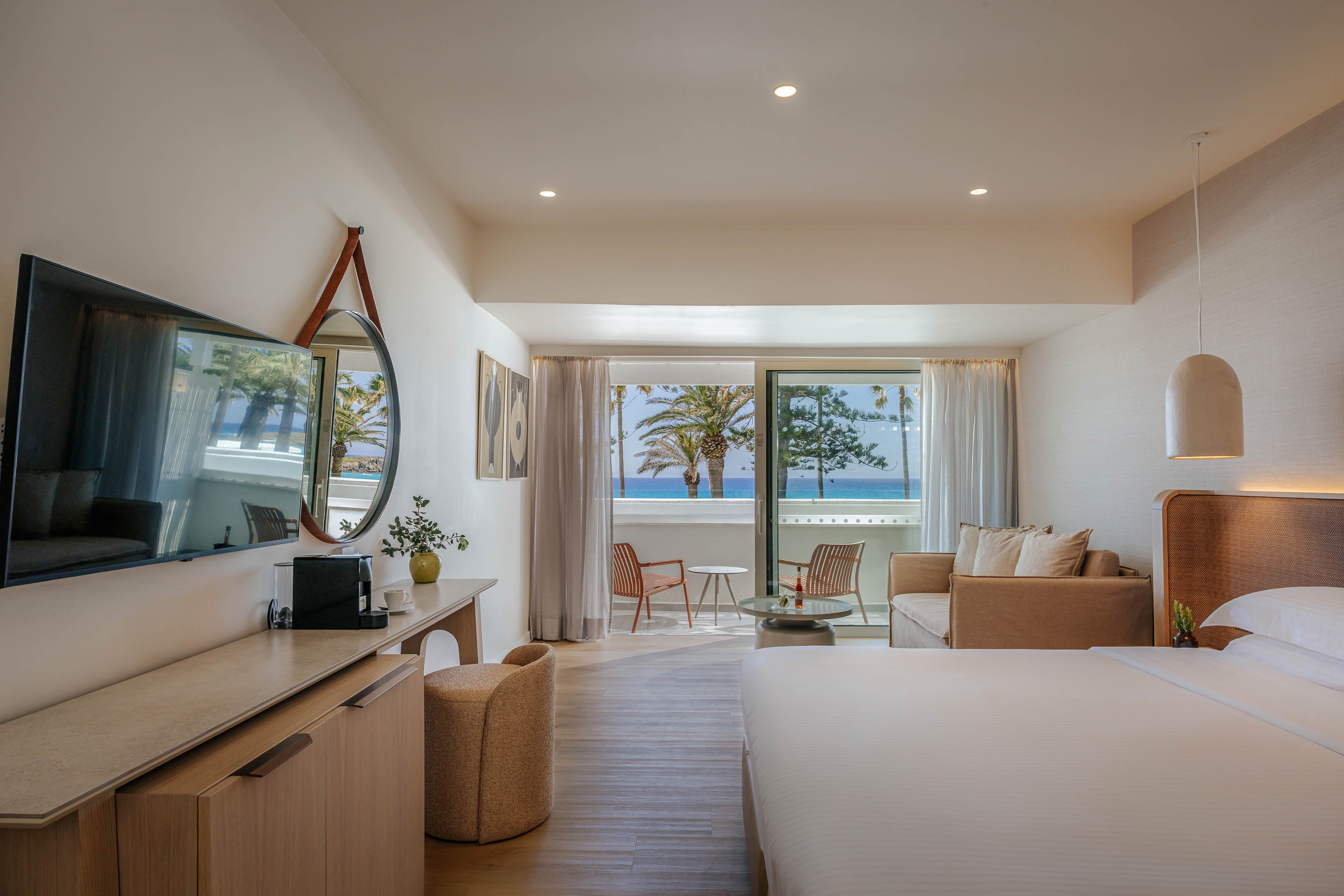 Contemporary hotel room featuring a large bed, sofa seating area, and wall-mounted flat-screen TV. Light wood furniture and neutral tones create a calm, minimalist atmosphere. A balcony with chairs overlooks palm trees and the ocean, visible through wide glass doors. Soft lighting, decorative artwork, and a round mirror add warmth and style to the coastal interior.
