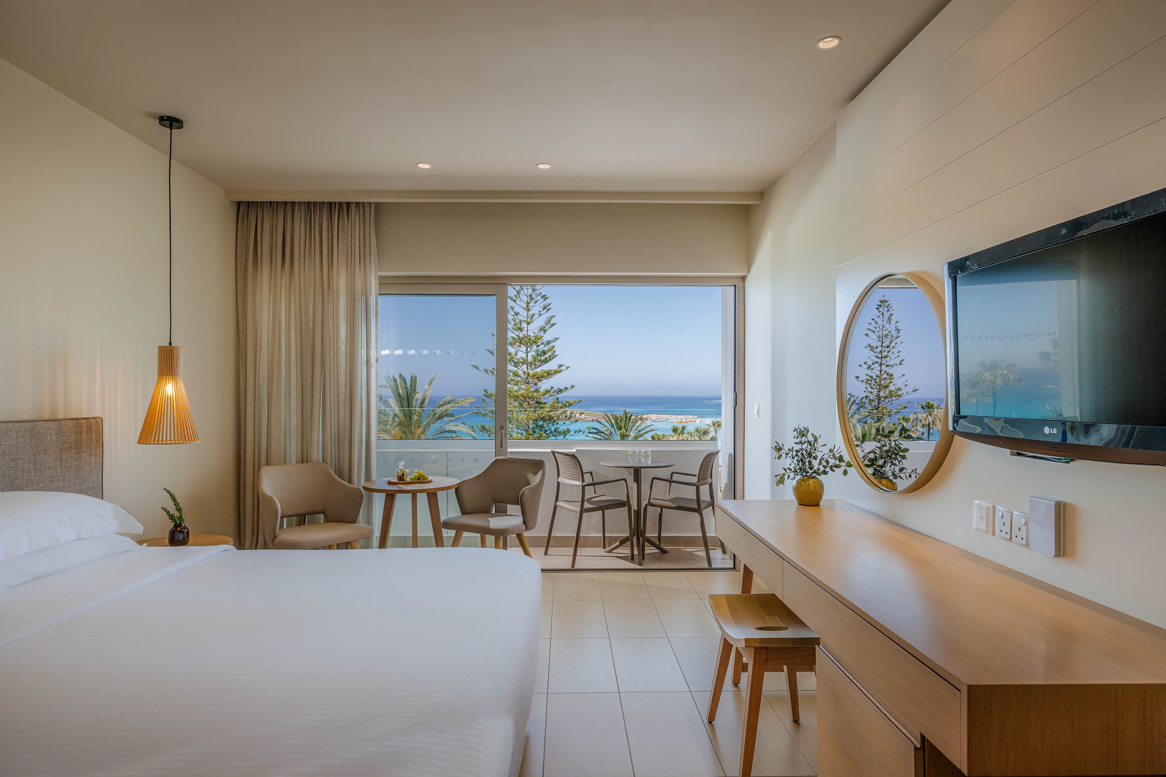 A contemporary hotel room features a neatly made bed and a long wooden desk with a wall-mounted television. Soft neutral tones and warm lighting create a calm, inviting atmosphere. Near the window, a small seating area with chairs and a table leads out to a balcony overlooking palm trees and the ocean. Large glass doors frame the scenic coastal view, enhancing the sense of openness and relaxation.