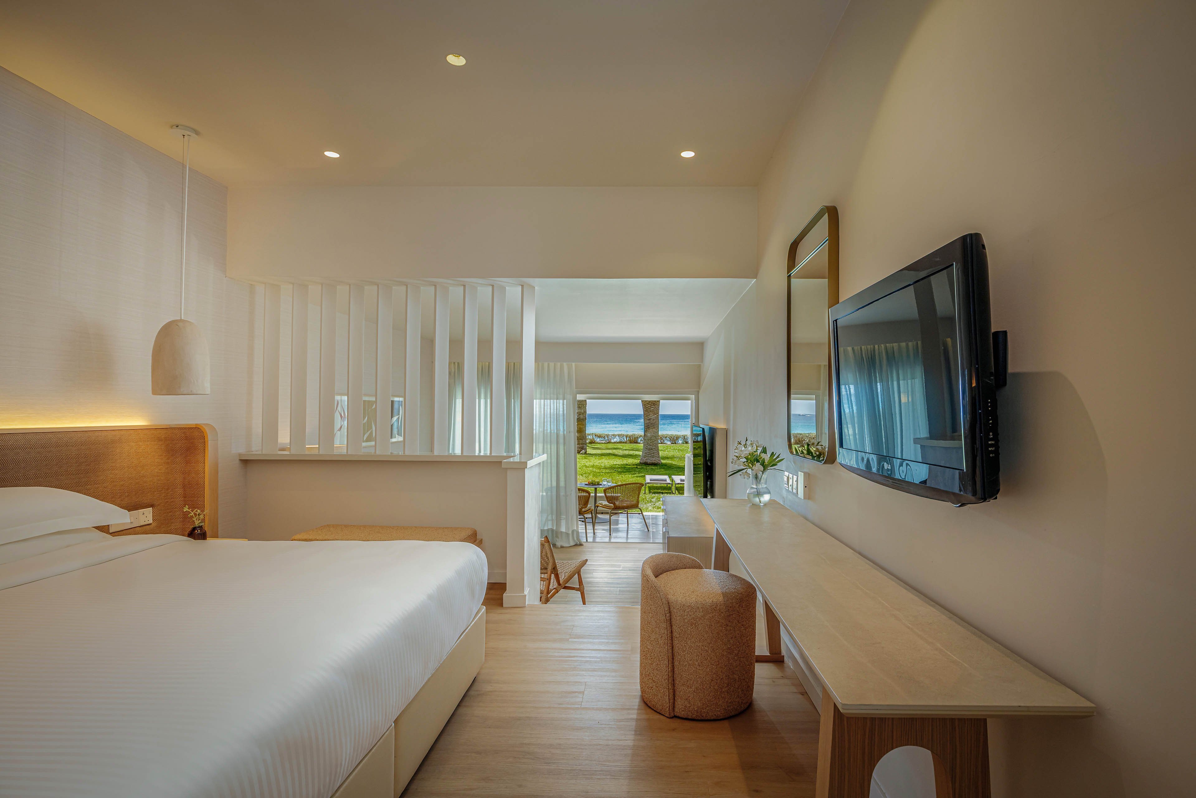 A contemporary hotel room features a large bed with crisp white linens and a minimalist wooden headboard. A flat-screen television is mounted on the wall opposite a long wooden desk with a round upholstered stool. Soft neutral tones and warm lighting create a calm, inviting atmosphere. At the far end of the room, sliding doors open to a balcony overlooking green lawns and the ocean beyond.