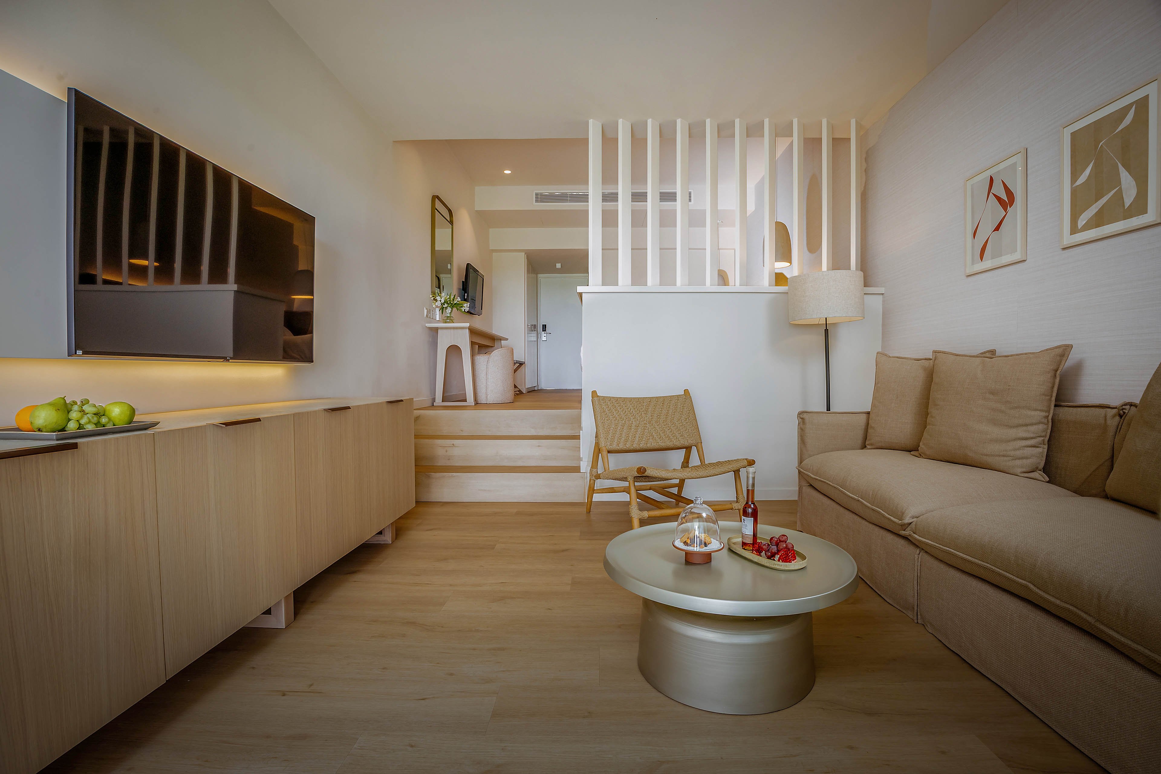 Contemporary living room interior featuring a neutral-toned sofa and a sleek wall-mounted television. A round coffee table with drinks and fruit sits in the center, while light wood flooring and cabinetry create a cohesive look. Vertical white slats separate the slightly elevated back area with a chair and desk, adding architectural interest. Soft, warm lighting and abstract artwork on the wall contribute to a calm, inviting atmosphere.