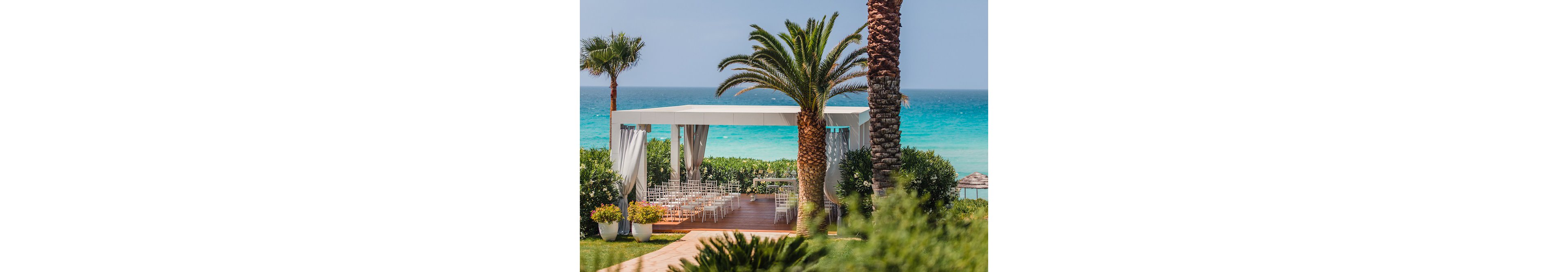 An elegant white pavilion is set up for a wedding ceremony overlooking a turquoise sea. Rows of white chairs are neatly arranged on a wooden deck under the canopy. Lush green shrubs and palm trees frame the view, leading the eye toward the water. The bright daylight and clear sky create a warm, romantic coastal atmosphere.