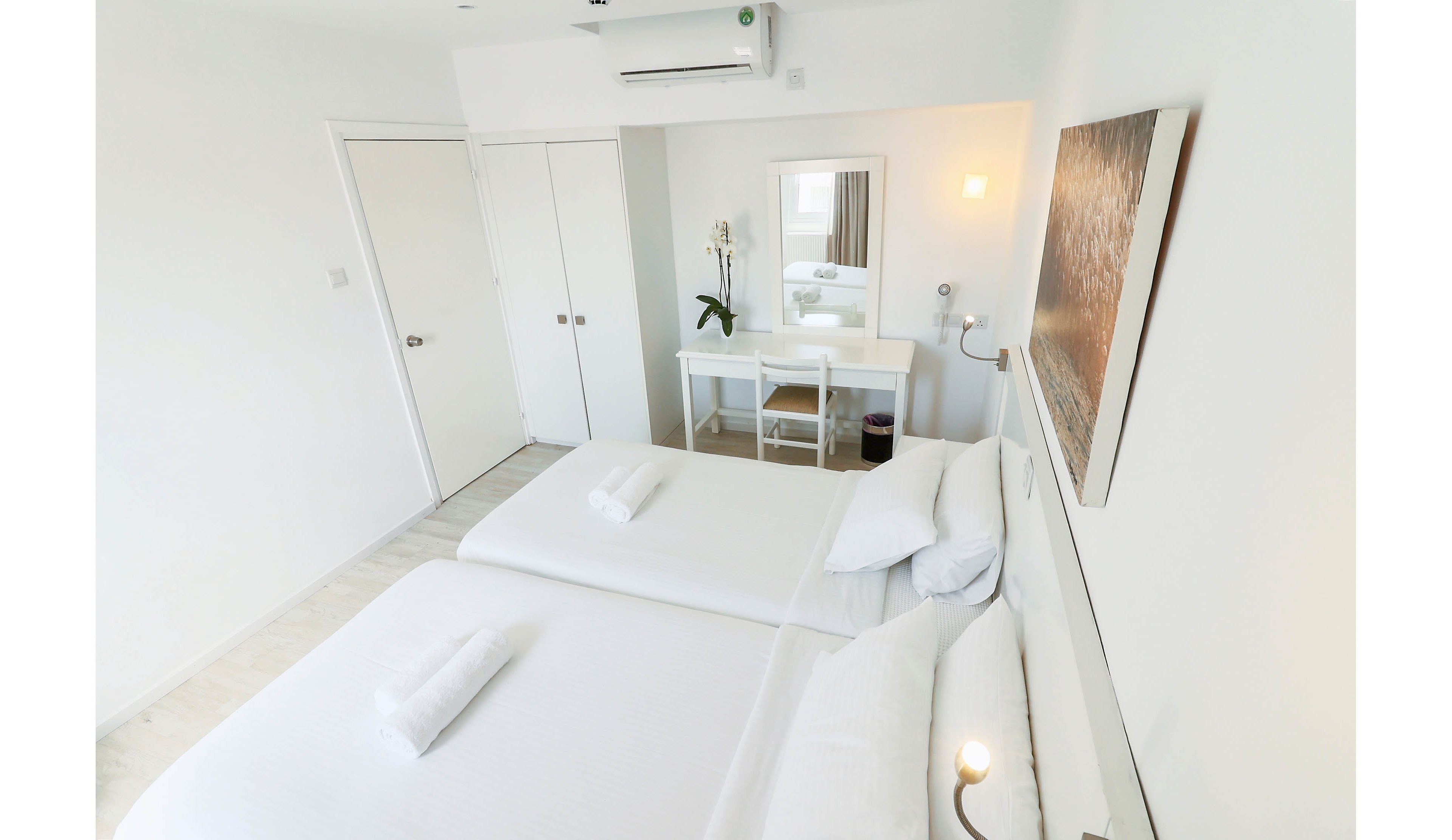 A bright, minimalist bedroom features two neatly made twin beds with white linens and rolled towels placed on top. The room is decorated in an all-white palette, creating a clean and modern atmosphere. A small desk with a mirror, chair, and potted orchid sits against the far wall, while an air conditioning unit is mounted above. Soft lighting and simple artwork contribute to the calm, contemporary feel of the space.
