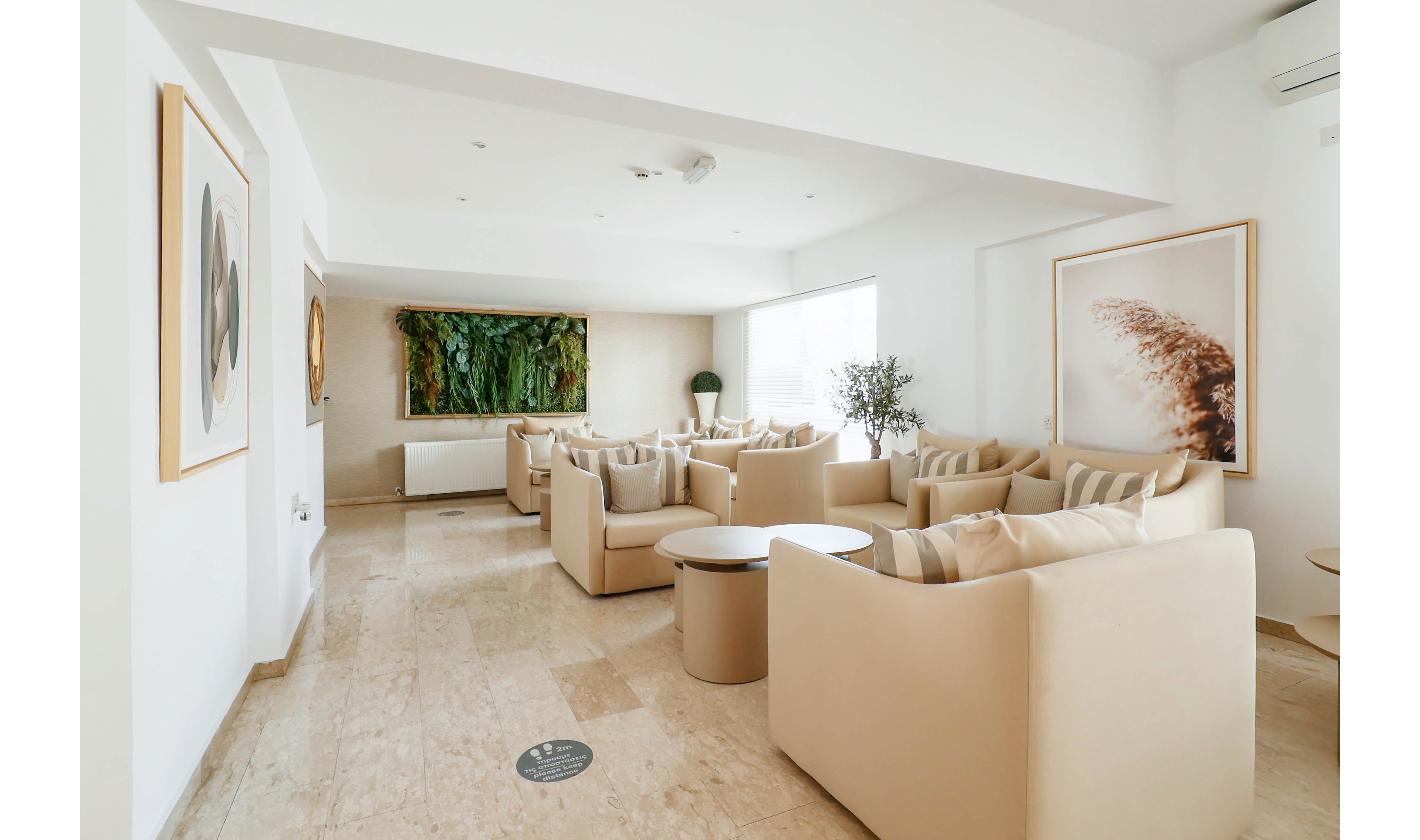 A contemporary lounge area features multiple beige armchairs arranged in conversational clusters. The space has a light, neutral color palette with marble or stone flooring and white walls. Large framed artwork, including a green vertical garden piece and an abstract print, decorates the walls. Natural light filters through sheer curtains, creating a calm and inviting atmosphere suitable for waiting or relaxing.