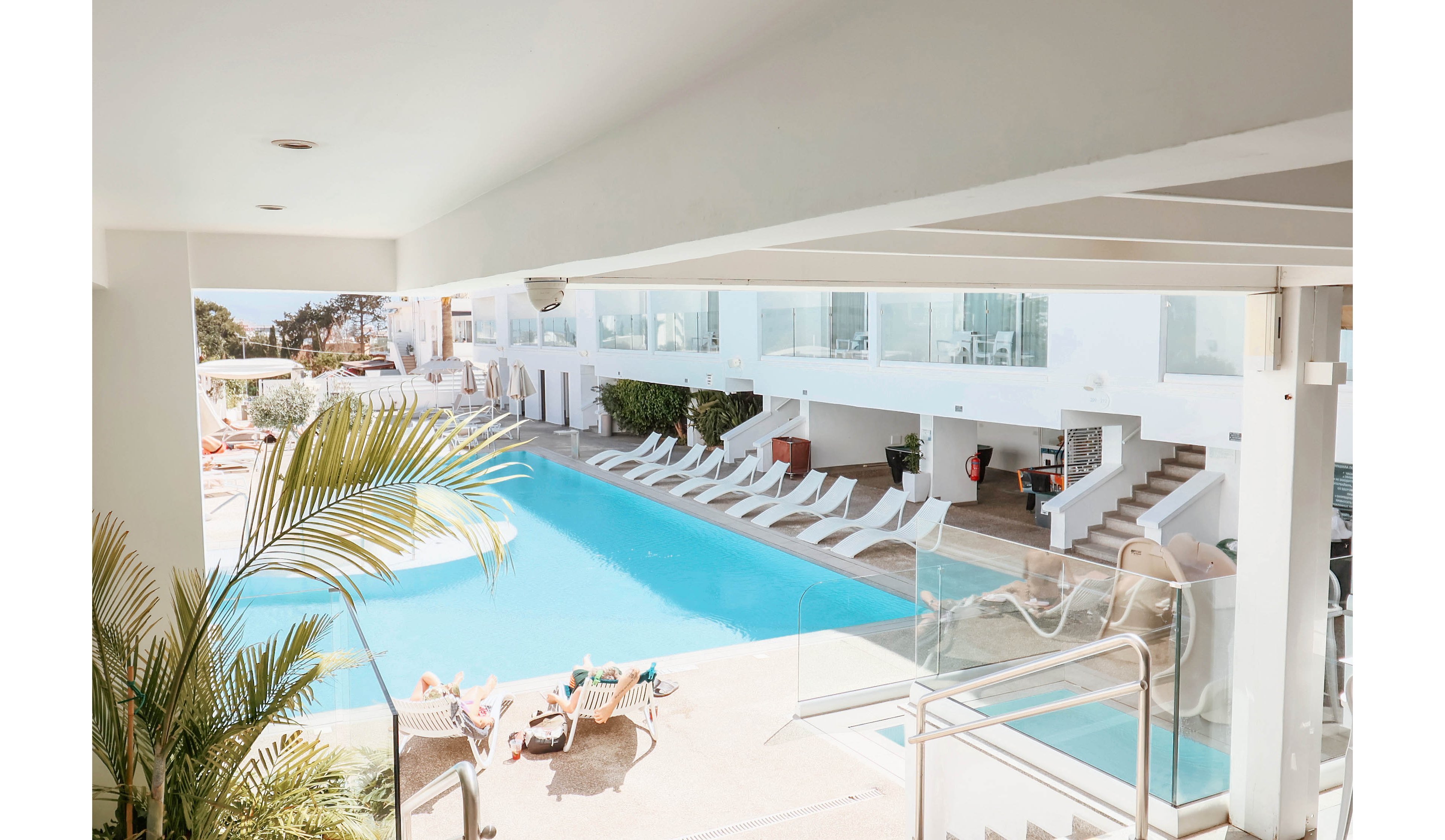 A bright, modern hotel pool area is shown under clear daylight. White lounge chairs line the turquoise swimming pool, with a few guests sunbathing near the water. Glass railings, clean architectural lines, and stairways emphasize a contemporary resort design. Palm leaves in the foreground add a tropical touch and enhance the vacation atmosphere.