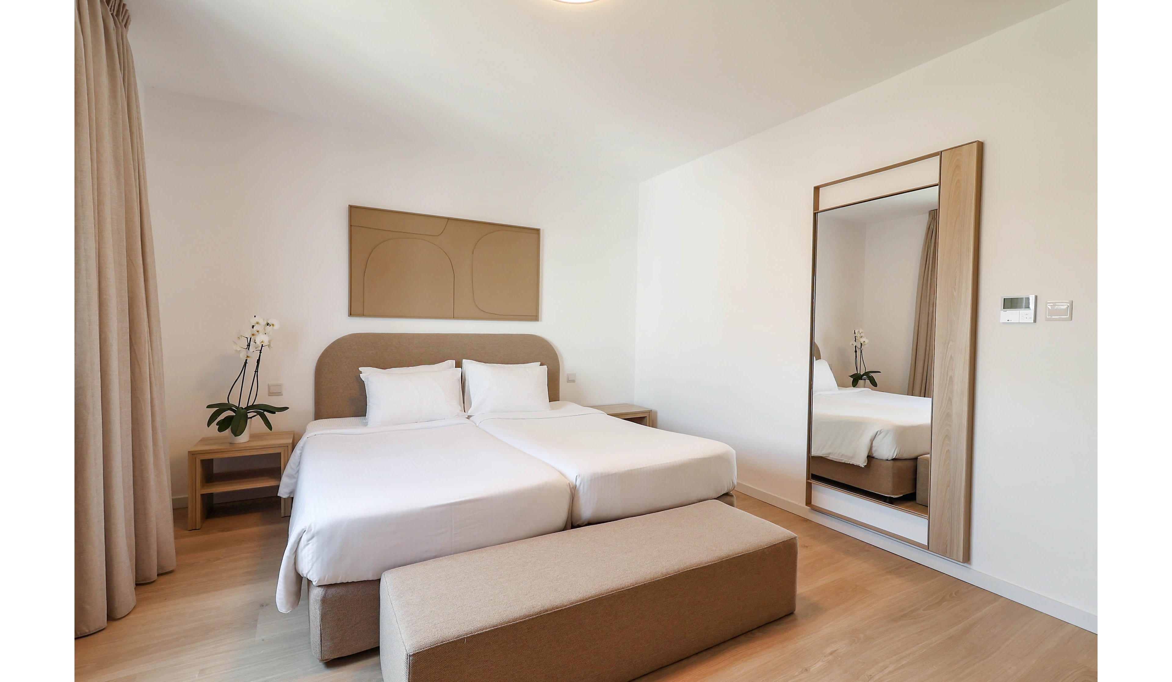 A bright, modern bedroom features two neatly made twin beds pushed together with crisp white linens. The room has a minimalist design with light wood flooring, a padded headboard, and a simple abstract wall art piece above the beds. A large floor mirror reflects the space, while a small bedside table holds a potted orchid. Neutral tones and soft natural light create a calm, contemporary atmosphere.