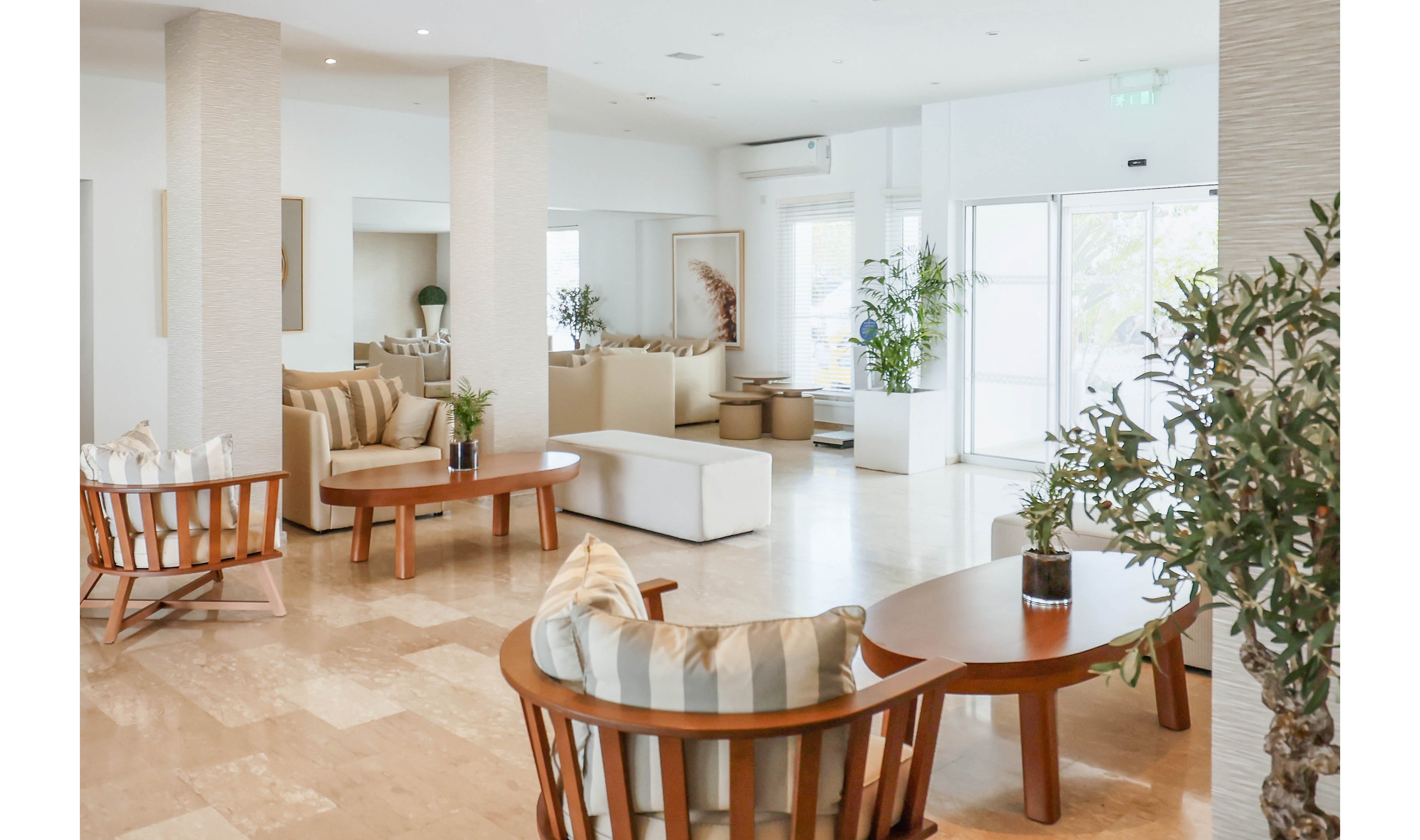 A bright, modern lounge area features wooden chairs and round tables arranged on a polished stone floor. Neutral-toned sofas with striped cushions create a comfortable seating space near tall white columns. Large windows and glass doors allow natural light to fill the room, highlighting potted plants and minimalist decor. The overall atmosphere is airy, clean, and welcoming, suitable for a hotel lobby or contemporary reception area.