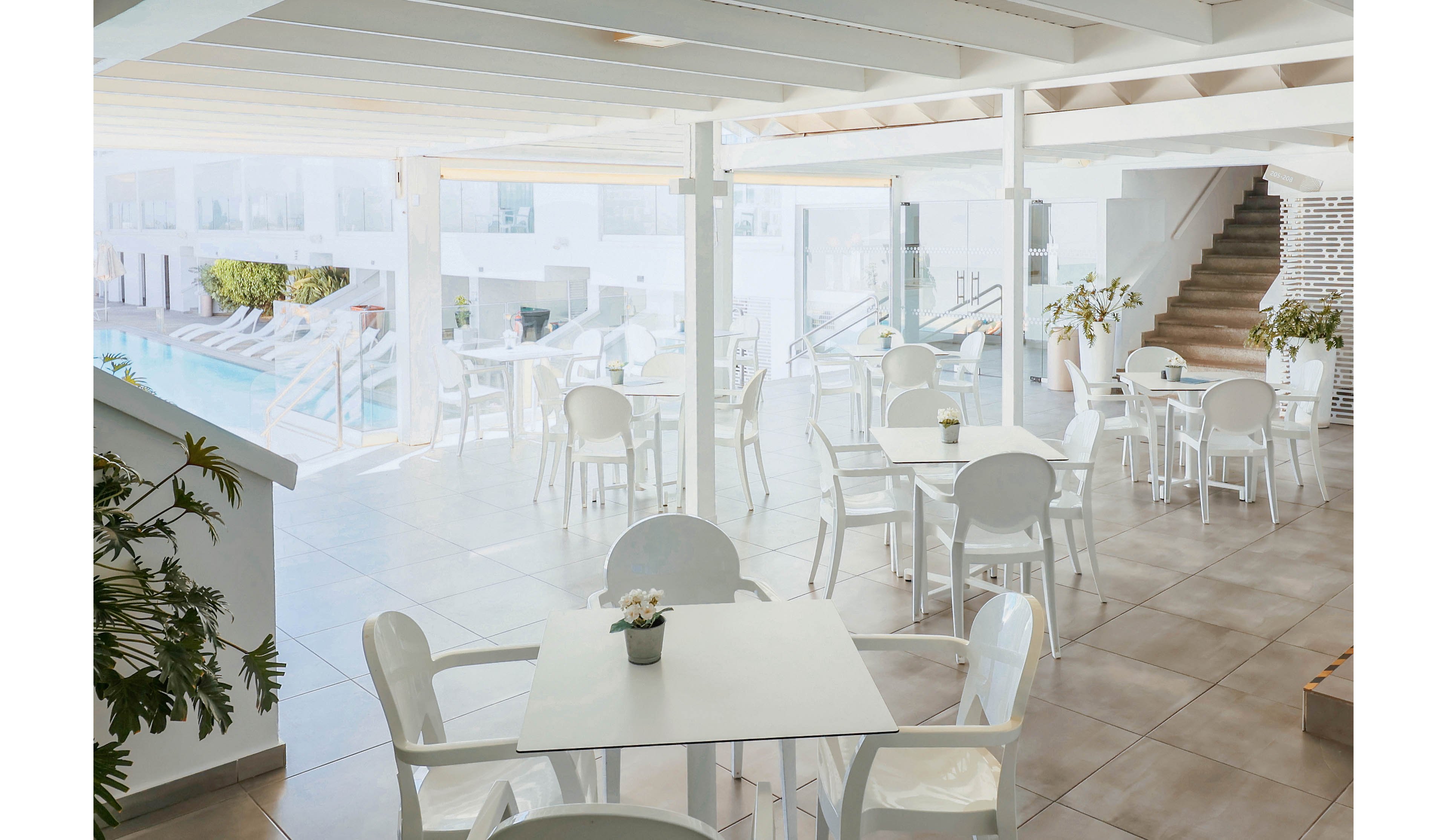 An airy poolside cafe features white tables and chairs arranged on a tiled terrace. Large windows and open spaces allow natural light to flood the area, creating a clean and minimalist atmosphere. Potted plants and greenery add subtle decorative touches around the seating. In the background, a swimming pool and steps suggest a relaxed resort or vacation setting.