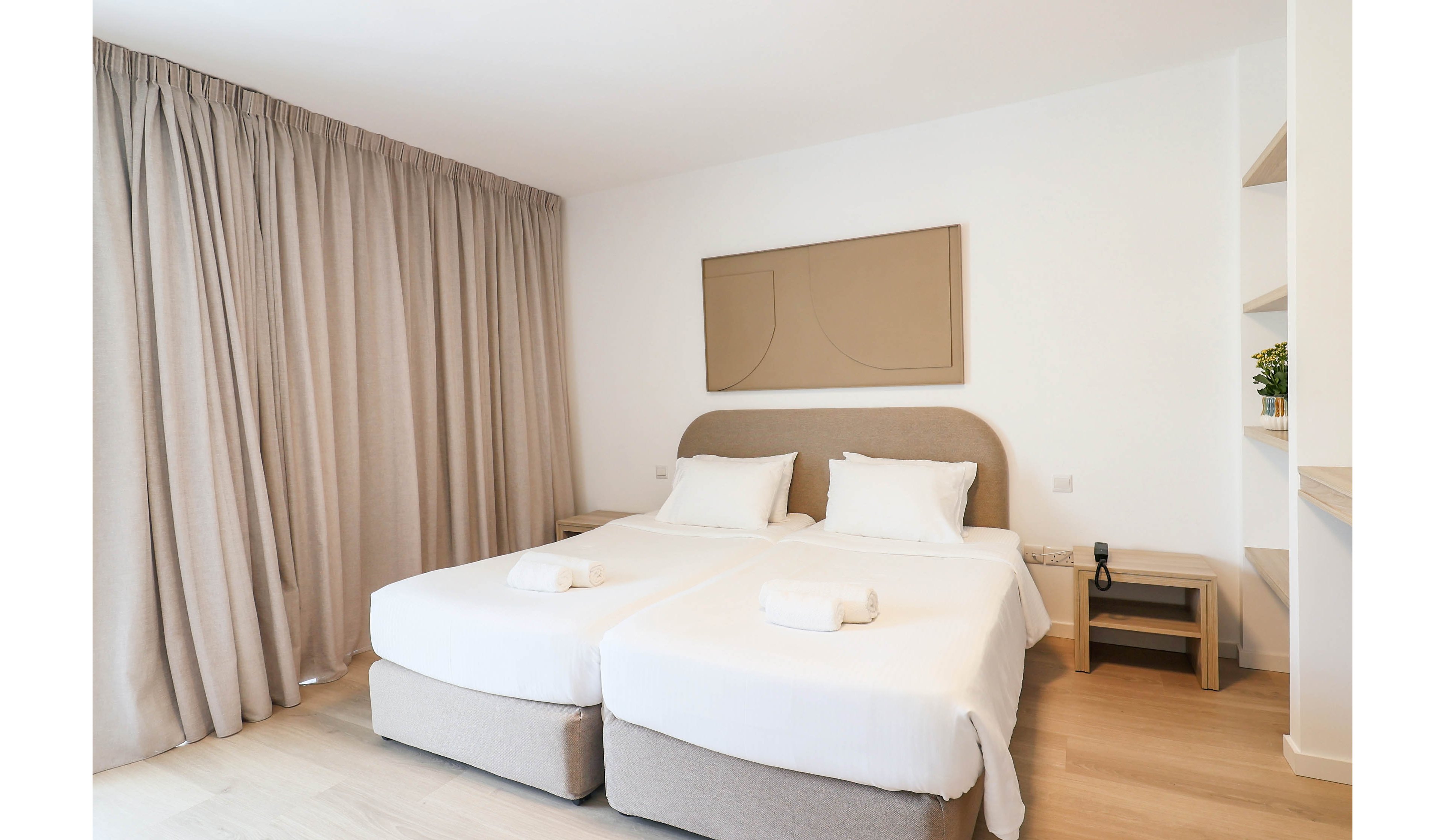 A bright, modern bedroom features two neatly made twin beds with white linens and folded towels placed on top. The room has light wood flooring, beige curtains covering a large window, and a minimalist wall art piece above the headboards. A small bedside table with a telephone and a built-in shelving unit with decorative plants add functional details. The neutral color palette and clean design create a calm, inviting atmosphere suitable for a hotel or guest room.