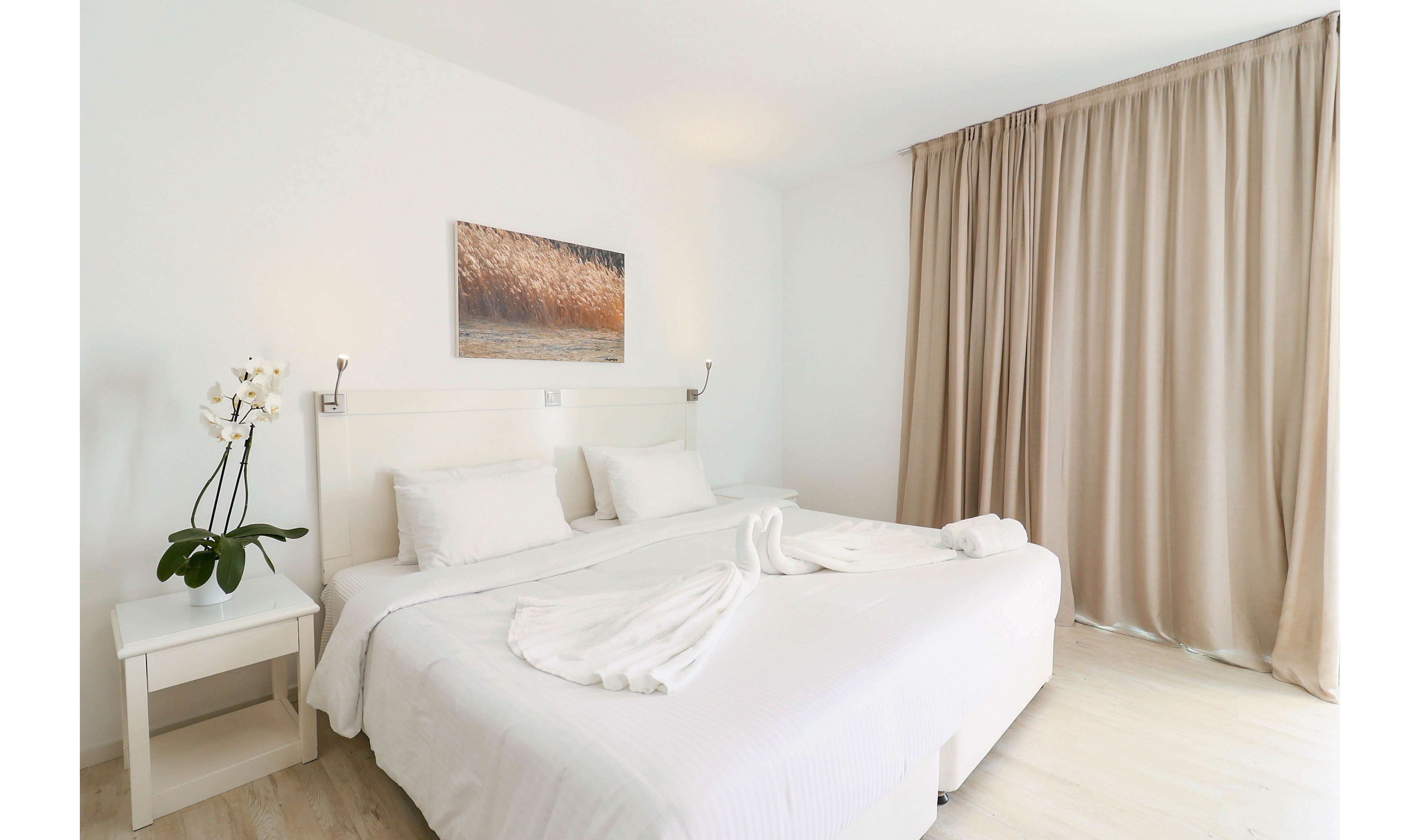 A bright, minimalist bedroom features two twin beds pushed together with crisp white linens and folded towel decorations. The room has light wooden flooring, a white headboard, and a small bedside table holding a potted orchid. Beige floor-to-ceiling curtains cover the window, adding warmth to the otherwise white interior. A framed landscape photograph hangs above the bed, completing the clean and modern hotel-style decor.