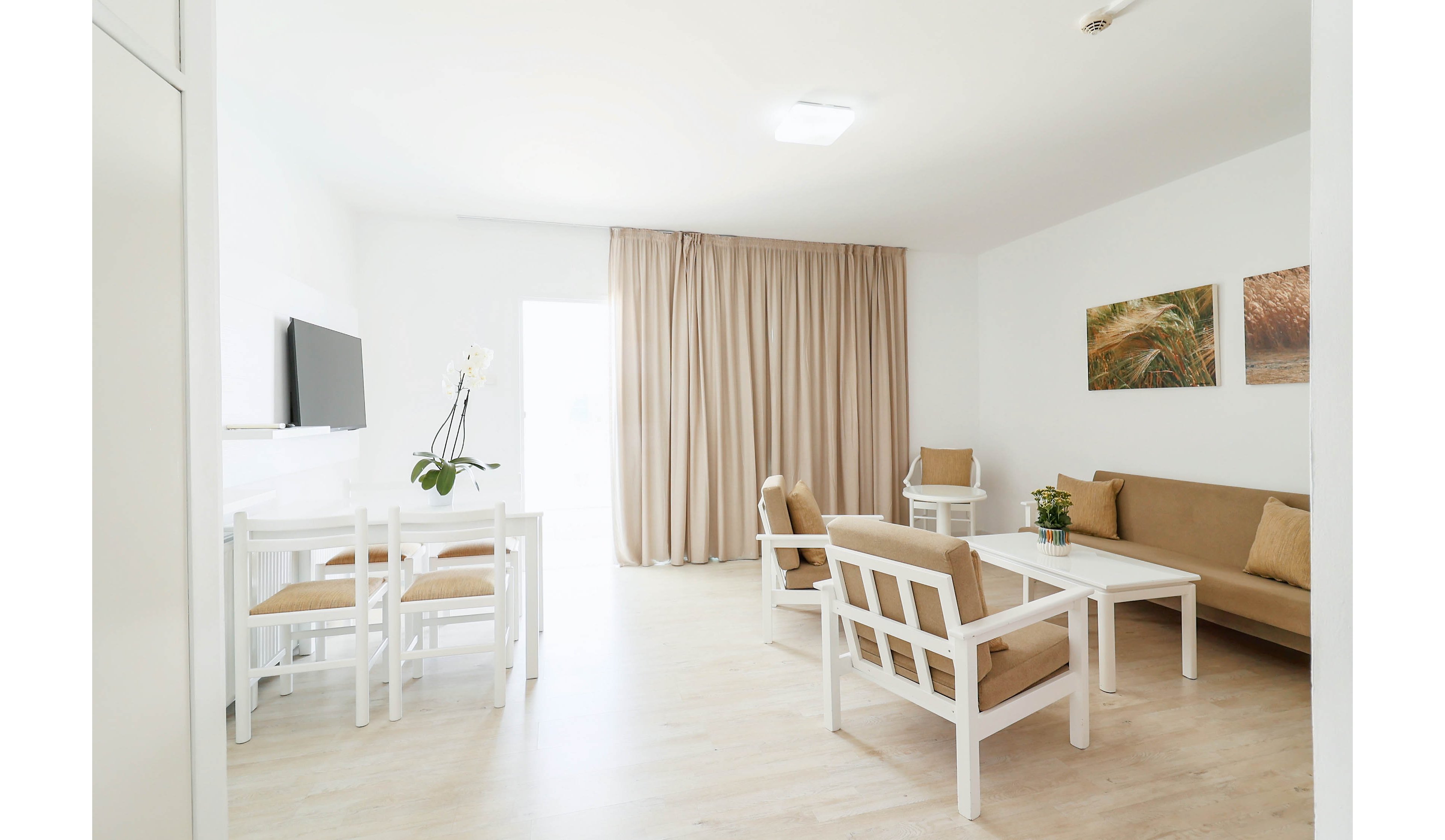 A bright, minimalist living room features white furniture and light wood flooring. A small dining table with four chairs sits near a wall-mounted television. A beige sofa and matching armchairs surround a low coffee table, with a potted plant adding a touch of greenery. Floor-to-ceiling beige curtains cover a large window or balcony door, softening the natural light. Two framed artworks hang on the wall, contributing subtle color and texture to the calm interior.