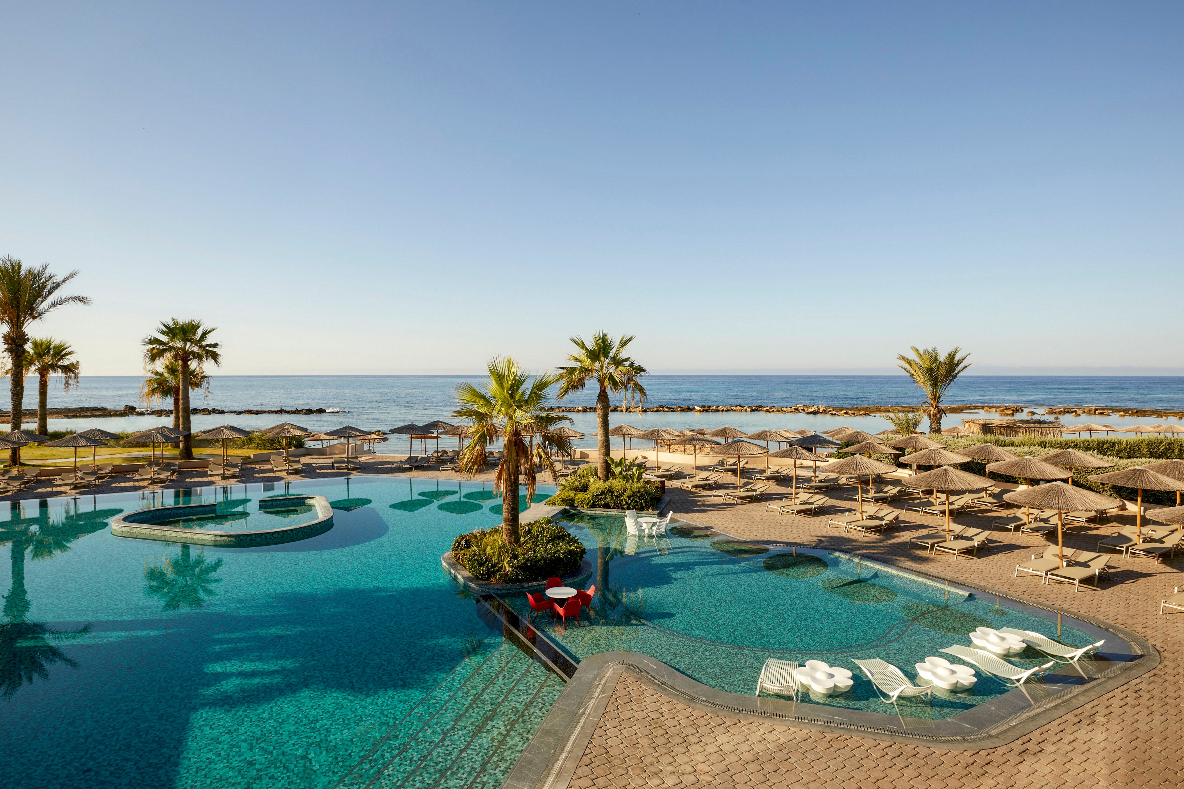 A large, tranquil swimming pool sits beside a sandy beach overlooking a calm sea. Palm trees and neatly arranged sun loungers with umbrellas create a relaxing vacation atmosphere. The clear blue water of the pool reflects the bright sky, while stone pathways and terraces frame the scene. In the distance, a rocky breakwater separates the open water from the resort shoreline.