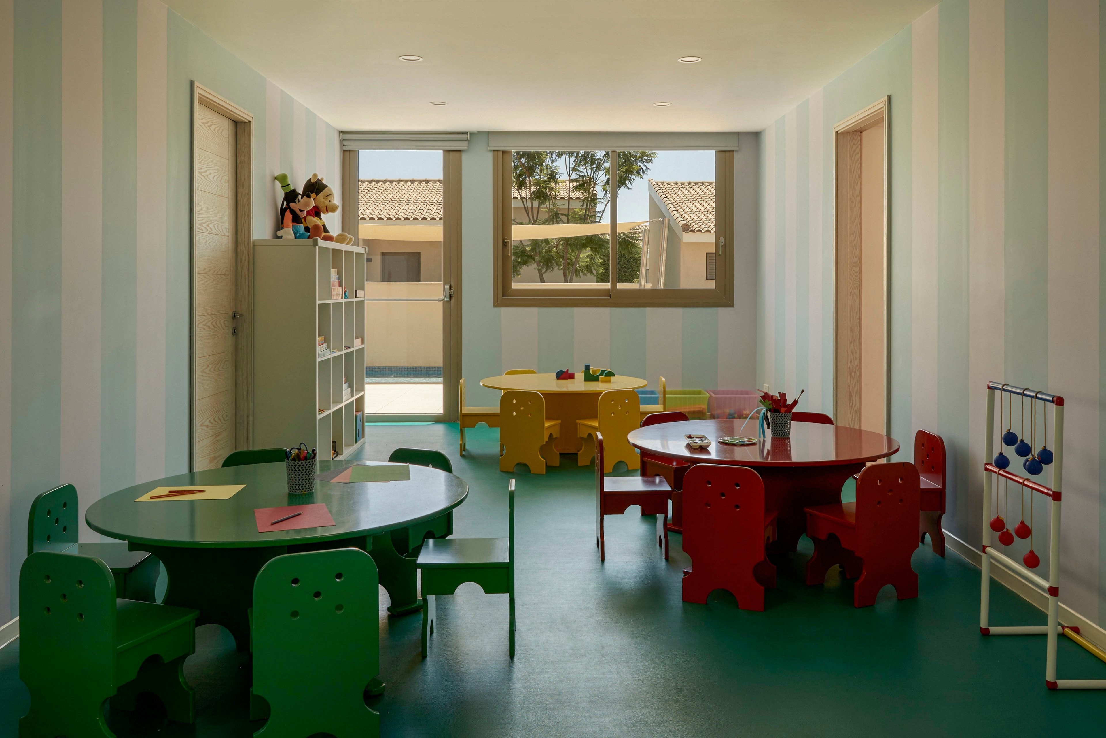 A brightly lit preschool classroom features several colorful round tables surrounded by small child-sized chairs. The room has soft pastel striped walls, teal flooring, and a large window overlooking an outdoor area. Shelving with toys and books stands against one wall, while simple educational toys are placed on the tables and in a corner. The overall atmosphere is cheerful and inviting, designed for young children’s play and learning.