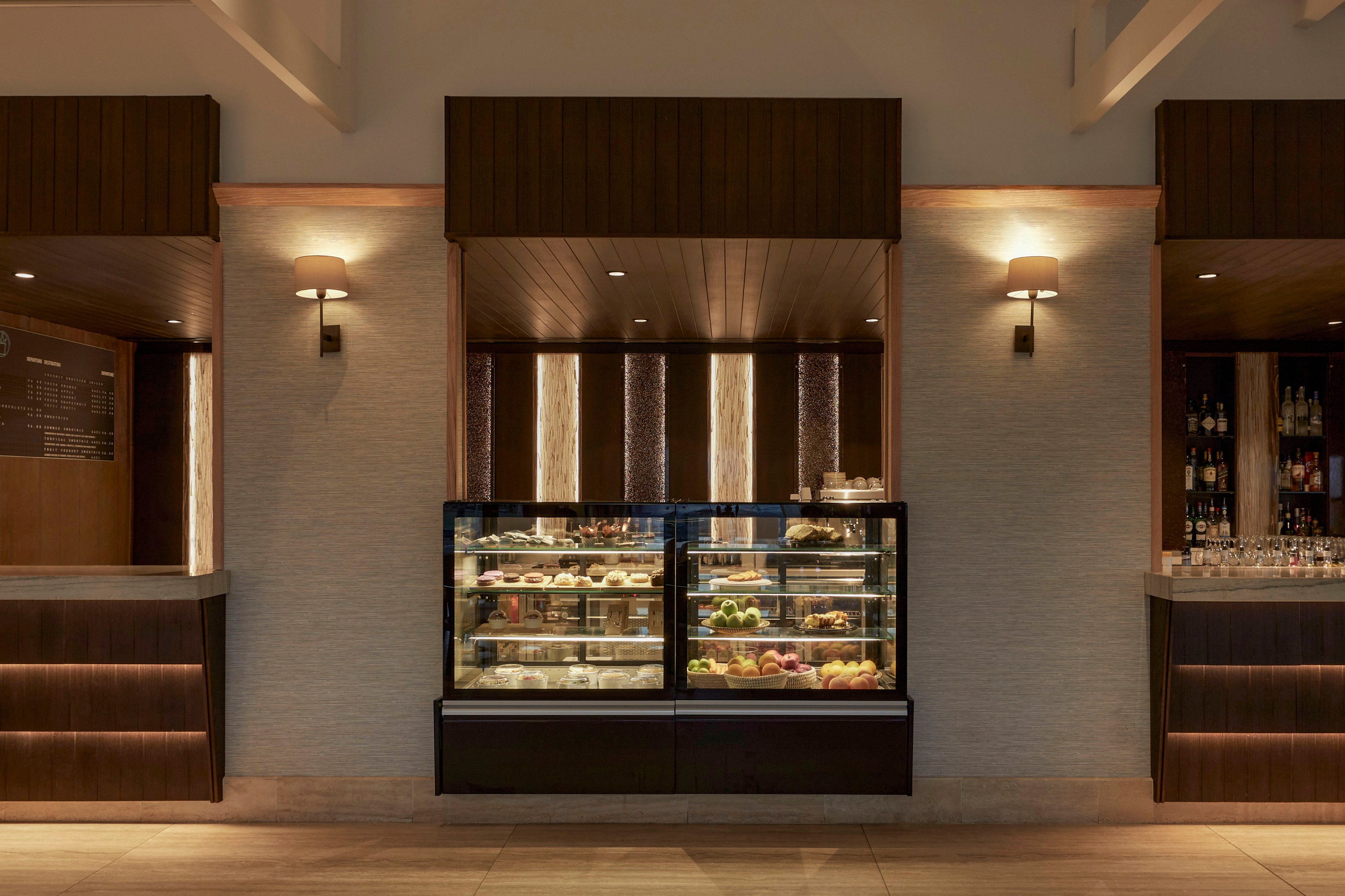 A contemporary café counter features a central refrigerated display case filled with pastries, desserts, and fresh fruit. The setting appears to be a hotel or upscale lobby bar, with warm wood paneling and soft wall sconces creating an inviting atmosphere. On the right side, shelves of bottles and glassware suggest a beverage or cocktail service area, while the left side includes a menu board. The overall scene is clean, symmetrical, and softly lit, emphasizing a refined, modern hospitality environment.