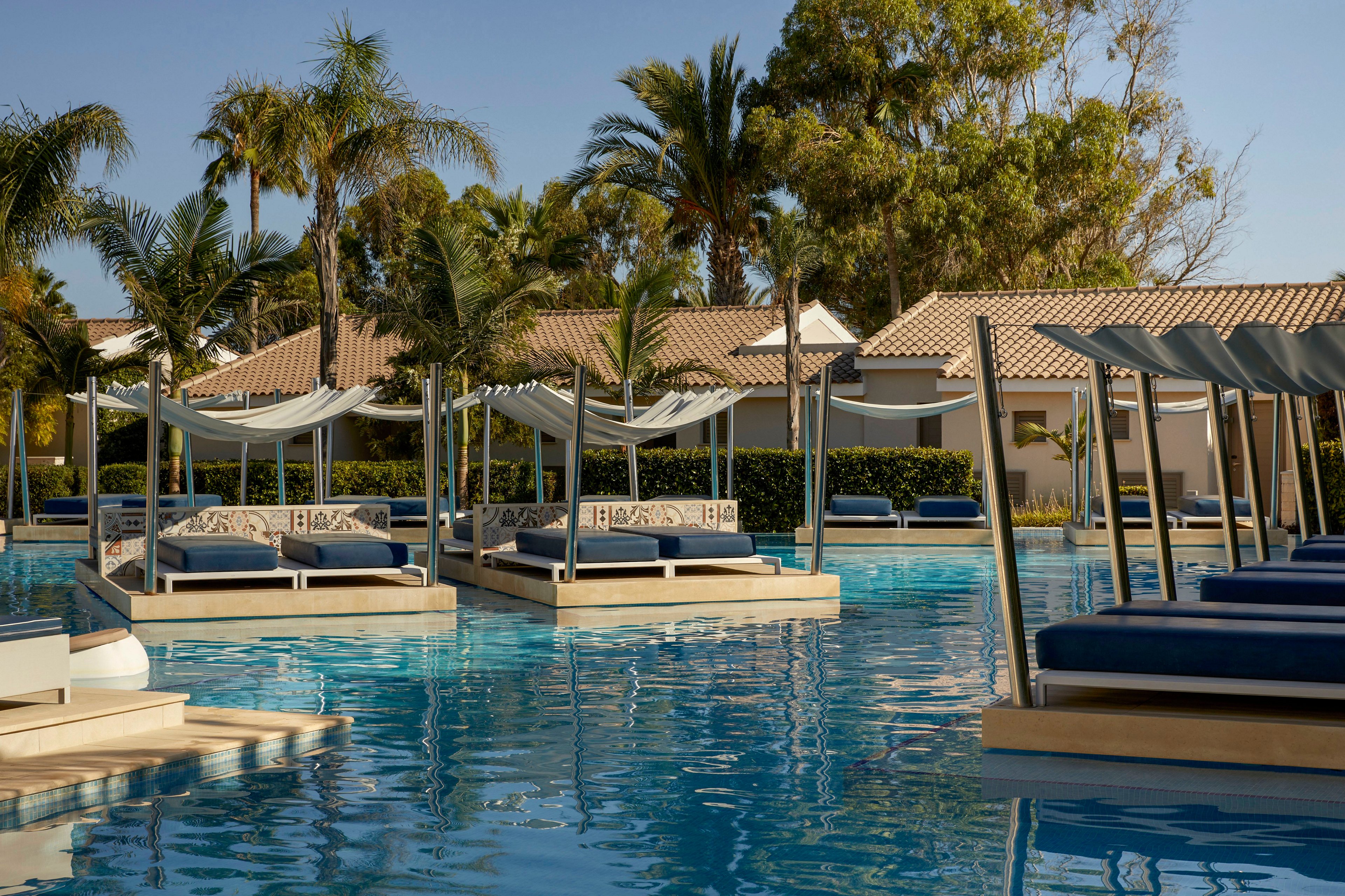 The image shows a serene resort swimming pool with multiple floating cabana-style daybeds. White fabric canopies drape over the beds, creating shaded lounging areas above the clear blue water. Palm trees and lush greenery surround the pool, with tiled-roof buildings in the background under a clear sky. The overall scene conveys a luxurious, relaxing vacation atmosphere in a warm, tropical setting.