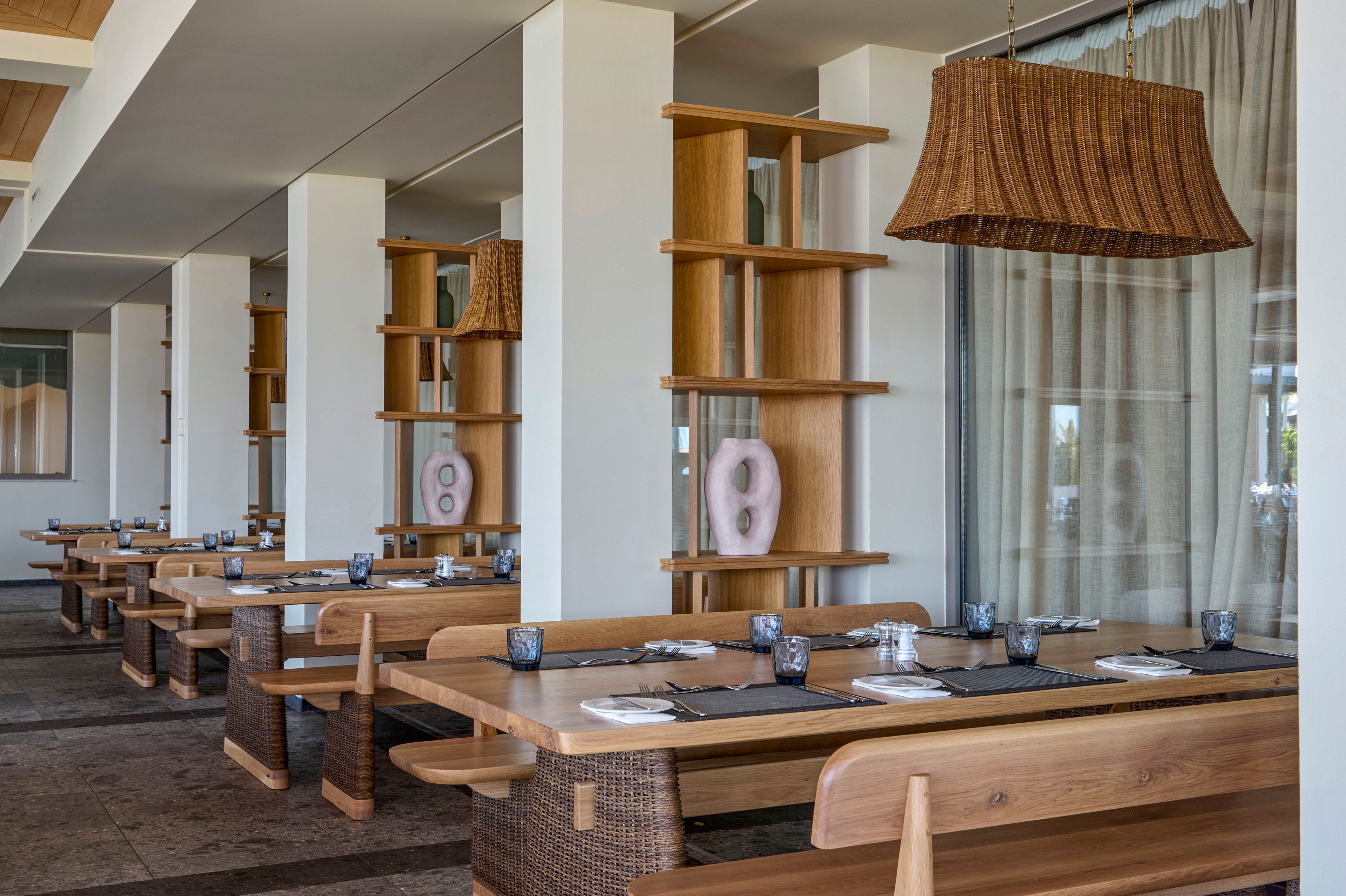 Contemporary restaurant dining area featuring long wooden tables and bench seating is shown in a clean, minimalist style. The setting includes woven chairs, wicker light fixtures, and open shelving with decorative vases along a row of columns. Neutral tones of wood, beige, and soft gray dominate the palette, creating a calm and inviting atmosphere. Table settings with placemats, glassware, and napkins suggest readiness for guests in a relaxed, upscale environment.