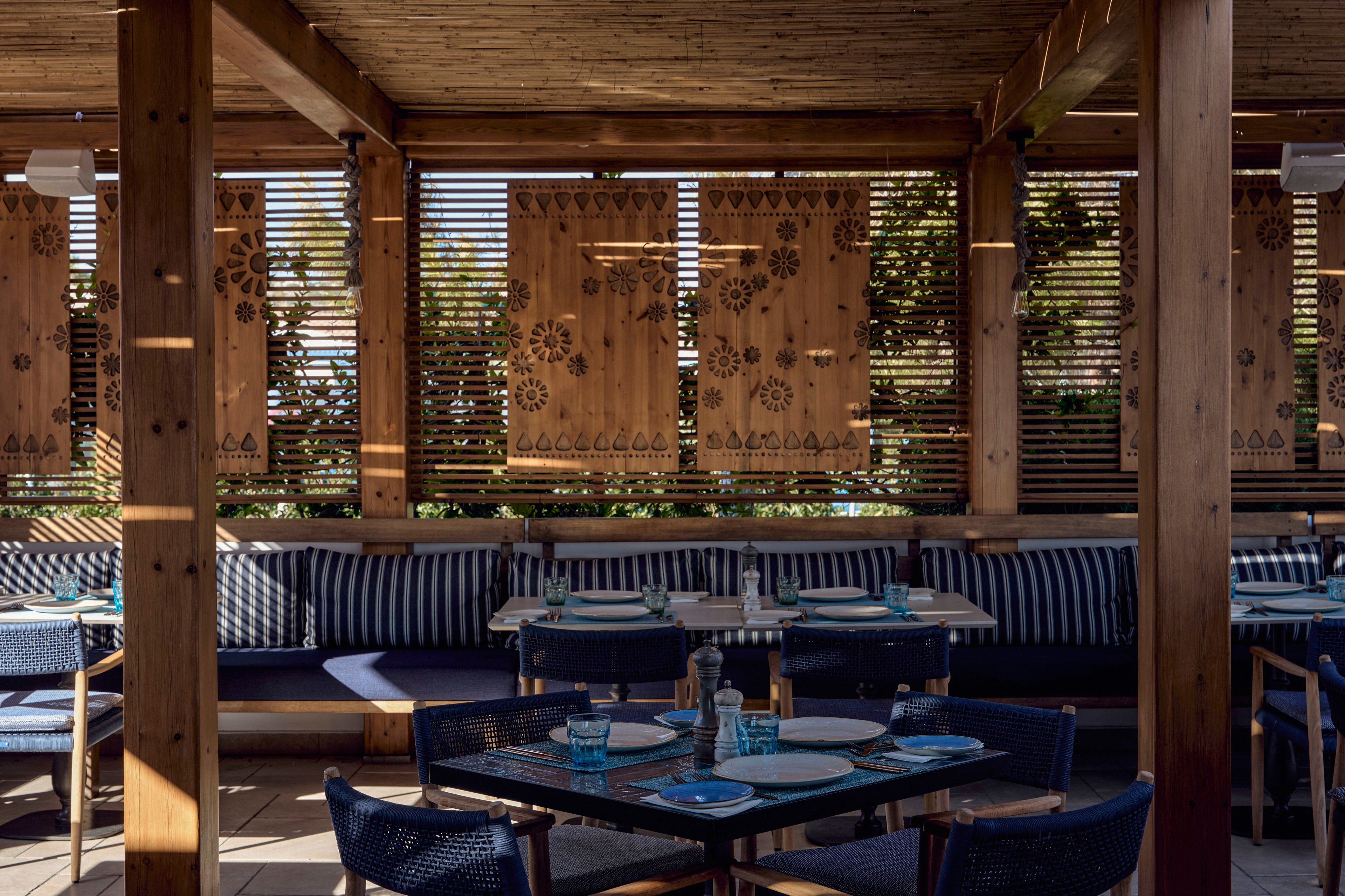 An outdoor restaurant terrace features wooden beams and decorative wooden panels with cutout patterns. Blue cushioned bench seating lines the perimeter, complemented by blue woven chairs around dark tables. Each table is neatly set with plates, glasses, and cutlery, suggesting readiness for guests. Sunlight filters through the slatted walls, creating a warm, relaxed atmosphere with natural and blue tones.