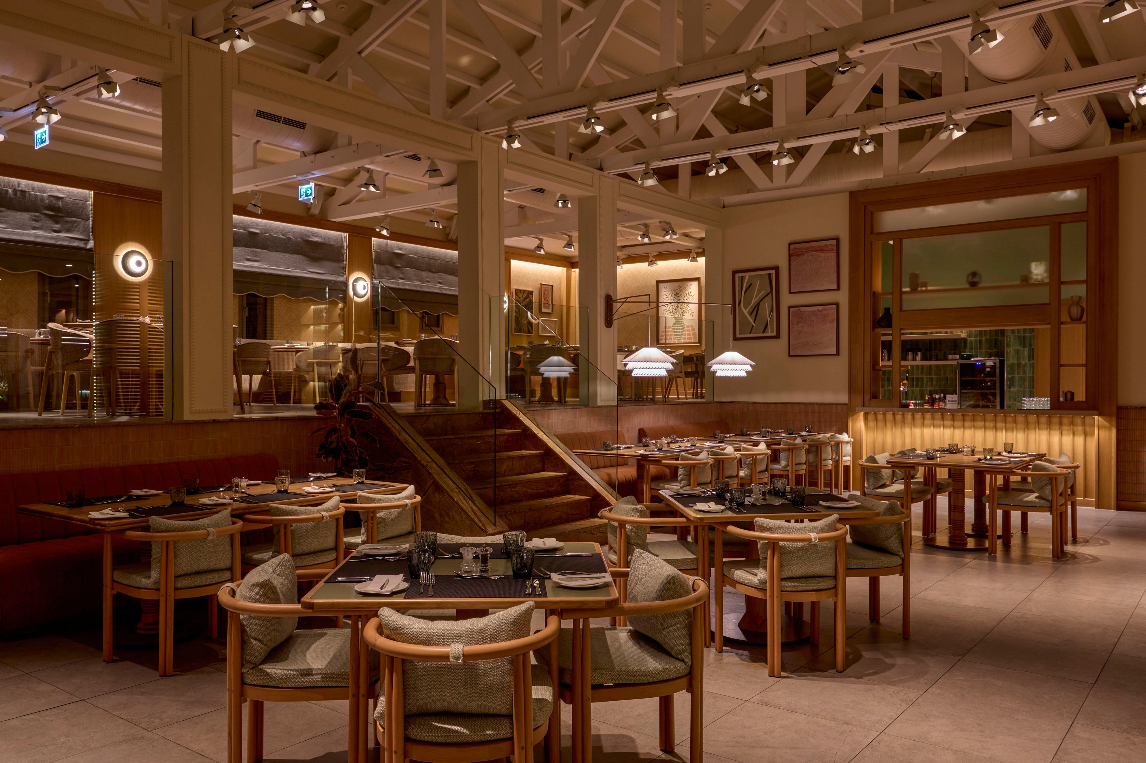 Upscale restaurant dining room with neatly arranged tables and chairs is shown in warm ambient lighting. The space features high ceilings with exposed beams, soft pendant lights, and wall sconces creating a cozy atmosphere. A central staircase leads to an elevated seating area, while framed artwork decorates the walls. The overall scene is calm and inviting, with no guests present and the tables prepared for service.