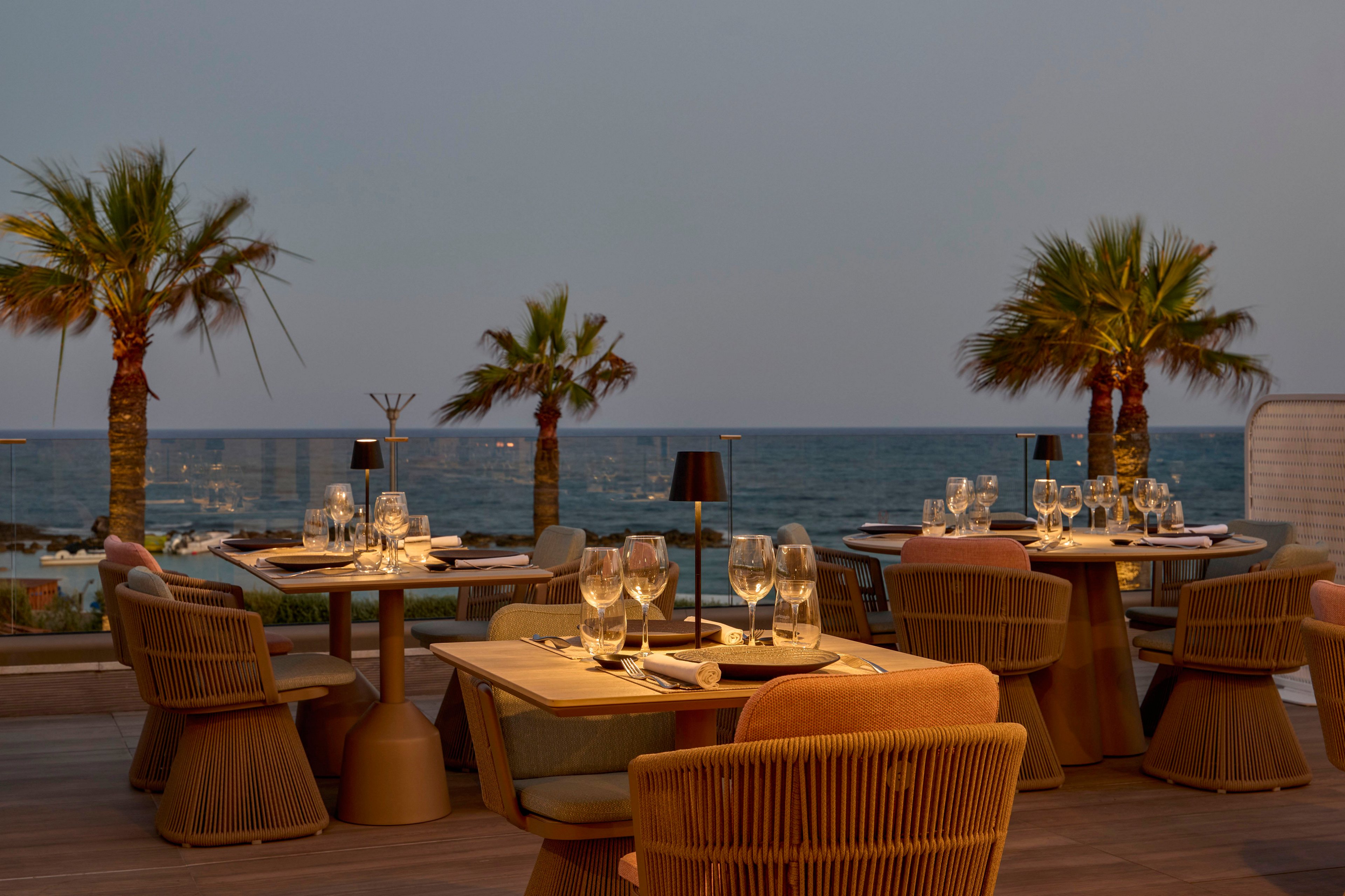 Outdoor restaurant tables are set with wine glasses and candles on a terrace overlooking the sea. Palm trees frame the view, and the soft evening light creates a warm, inviting atmosphere. Contemporary wicker chairs and neatly arranged table settings suggest a fine dining experience. The calm ocean and clear sky enhance the relaxed coastal ambiance.
