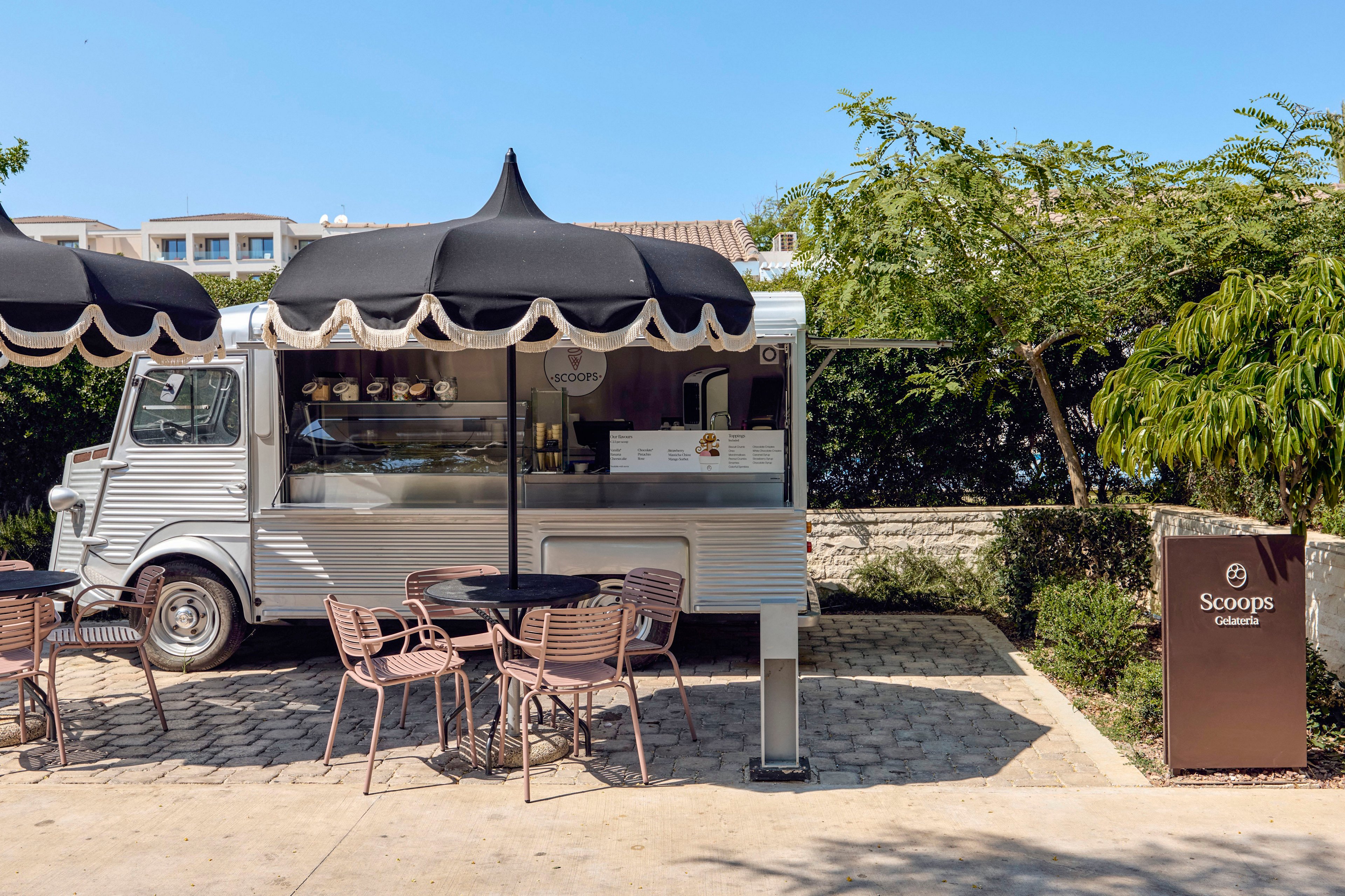 A silver food truck serves gelato under black-and-white scalloped awnings in an outdoor setting. Several empty metal café tables and chairs are arranged on a paved patio in front of the truck. A sign to the right clearly reads "Scoops Gelato" beside a simple graphic emblem. Lush greenery and distant buildings frame the relaxed, sunny atmosphere.
