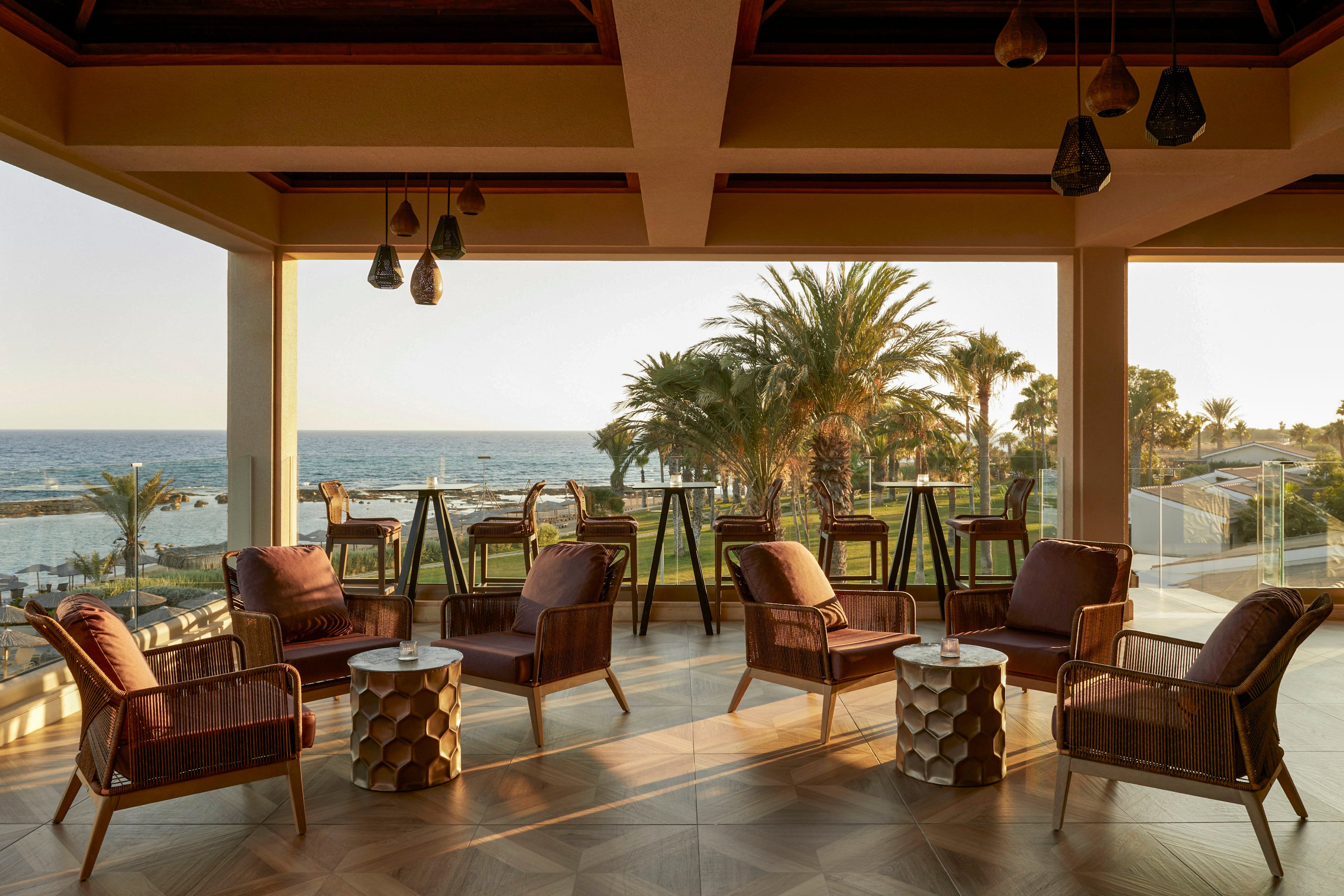An open-air lounge terrace overlooks the ocean, framed by large palm trees and a clear glass railing. Wicker armchairs with cushions are arranged around small geometric side tables on a patterned wooden floor. Warm sunset light fills the space, highlighting hanging pendant lamps and the relaxed seating area. The scene evokes a tranquil, upscale resort atmosphere ideal for evening relaxation.