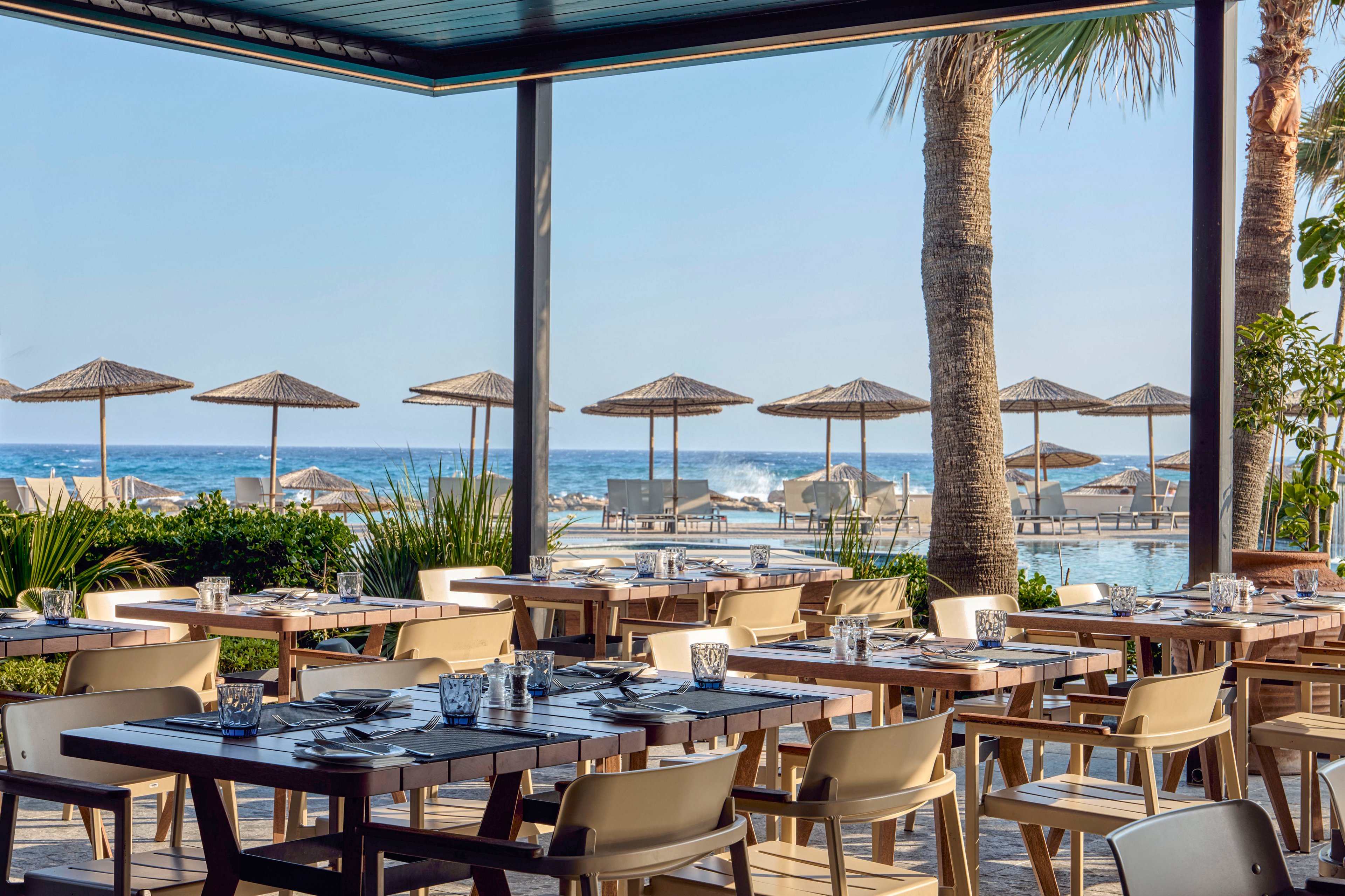 An open-air restaurant terrace overlooks a bright blue ocean and sandy beach. Wooden tables are neatly set with glassware, plates, and cutlery, surrounded by modern cream-colored chairs. In the background, straw sun umbrellas and lounge chairs line the waterfront near a pool area. Palm trees and lush greenery frame the scene, creating a relaxed vacation atmosphere under clear skies.