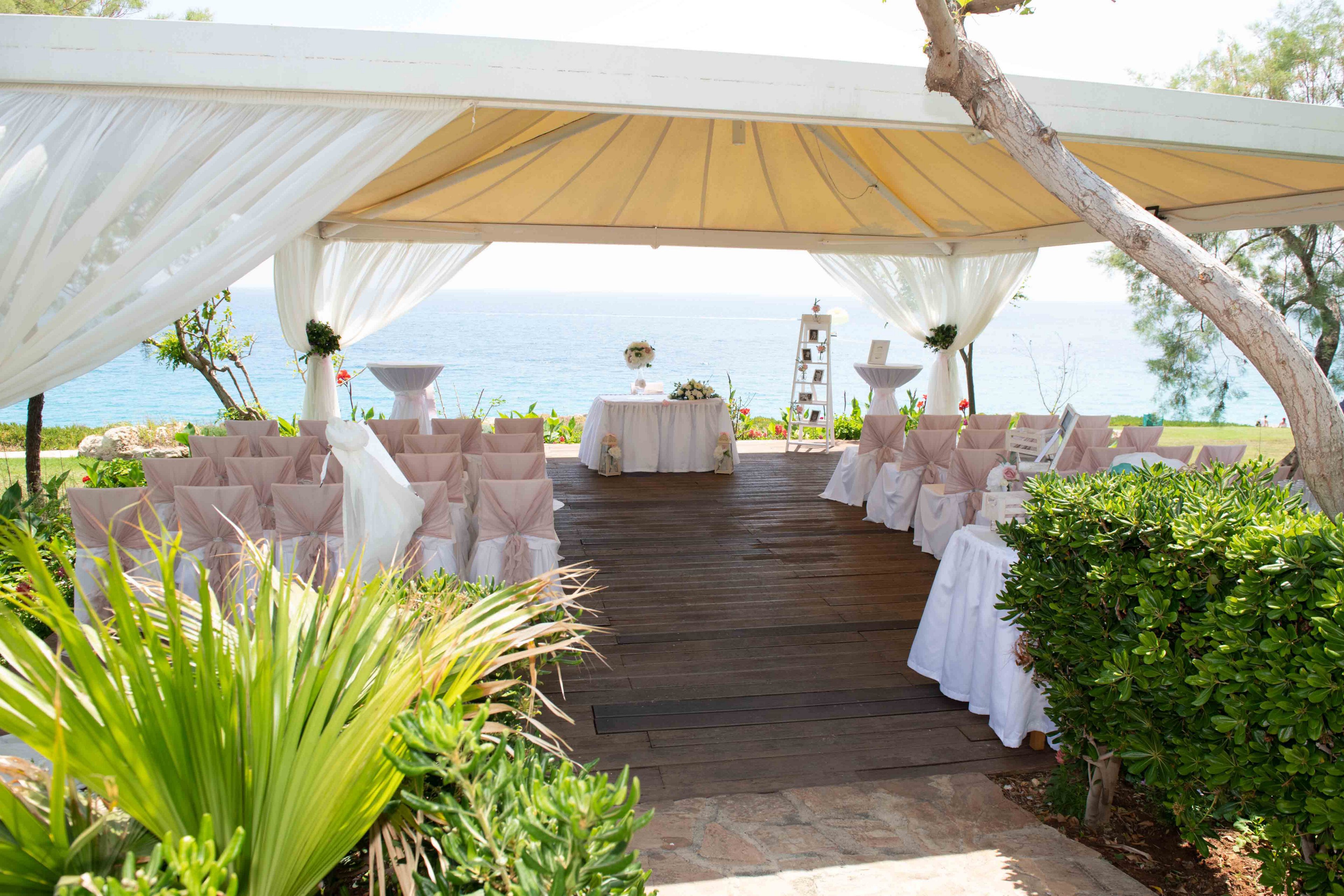 An elegant outdoor wedding ceremony venue is arranged under a large canopy overlooking the sea. Rows of chairs are neatly decorated with light fabric bows, facing a small table at the front. White draped curtains frame the open sides of the tent, allowing views of the blue water and sky. Lush green plants and shrubs surround the wooden aisle, adding a fresh, natural atmosphere to the romantic setting.