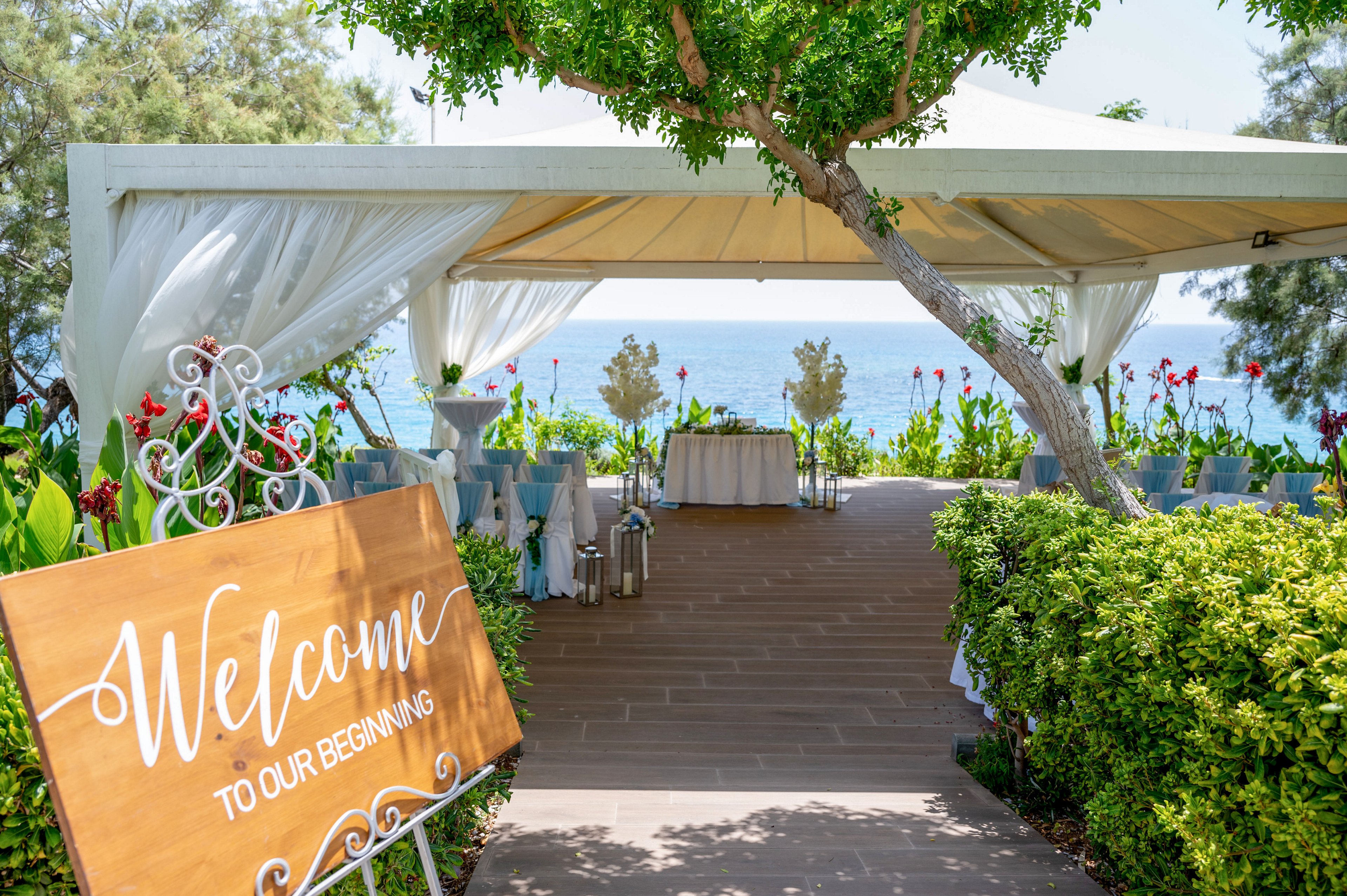 An elegant outdoor wedding reception entrance is set beneath a white canopy overlooking the sea. A large wooden sign reading "Welcome to our beginning" greets guests at the pathway. The venue is decorated with white drapes, blue chair sashes, floral arrangements, and lanterns. Lush greenery and ocean views create a romantic, summery atmosphere.