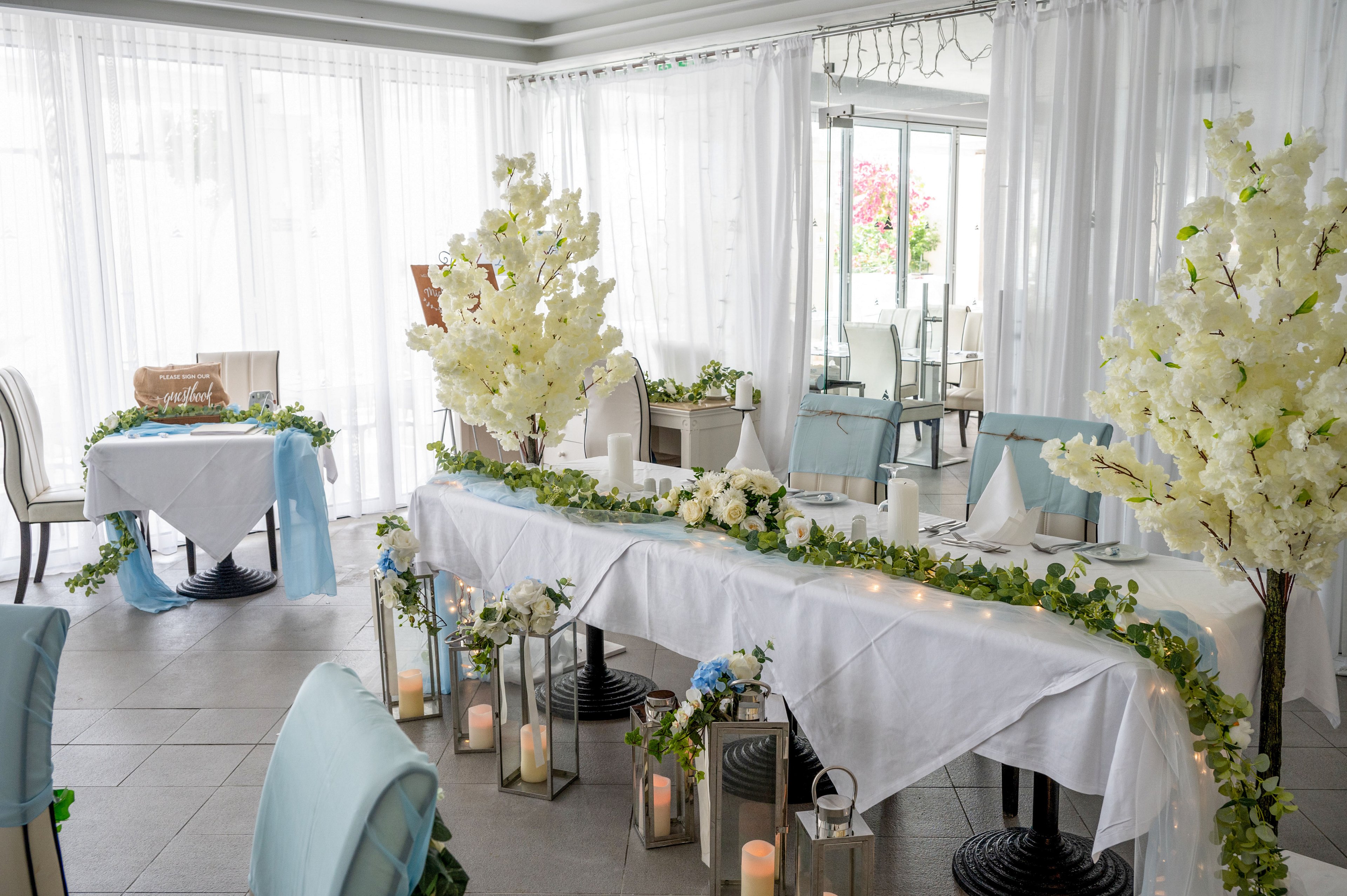 A long reception table is beautifully decorated with white linens, floral garlands, and soft greenery in a bright indoor venue. White blossom trees frame the scene, while lanterns with candles line the floor around the table. Light blue accents on chairs and floral details add a delicate touch to the overall arrangement. Sheer curtains and large windows create an airy, romantic atmosphere suitable for a wedding or formal celebration.