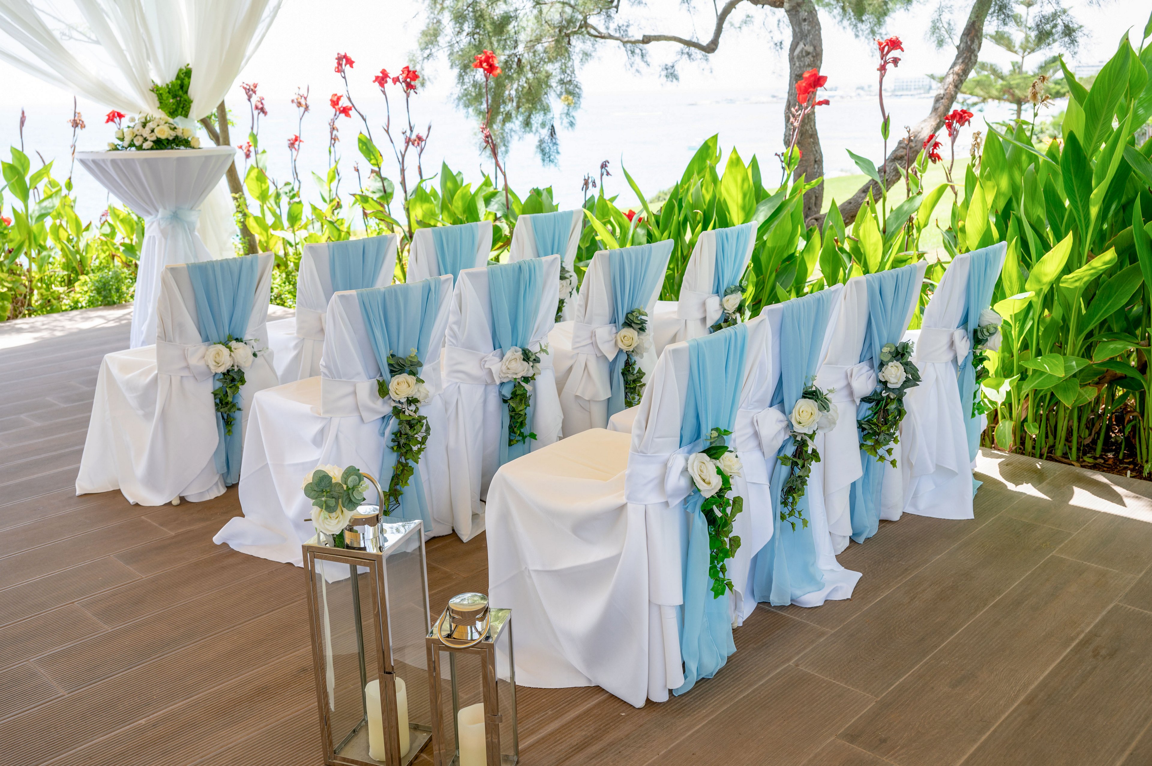 Rows of white chairs with light blue sashes are arranged for an outdoor wedding ceremony. Each chair is decorated with white flowers and green foliage, facing a small table draped in white fabric. Tall lanterns with candles stand along the aisle on a wooden floor. Lush green plants and red flowers frame the setting, with trees and water visible in the background.