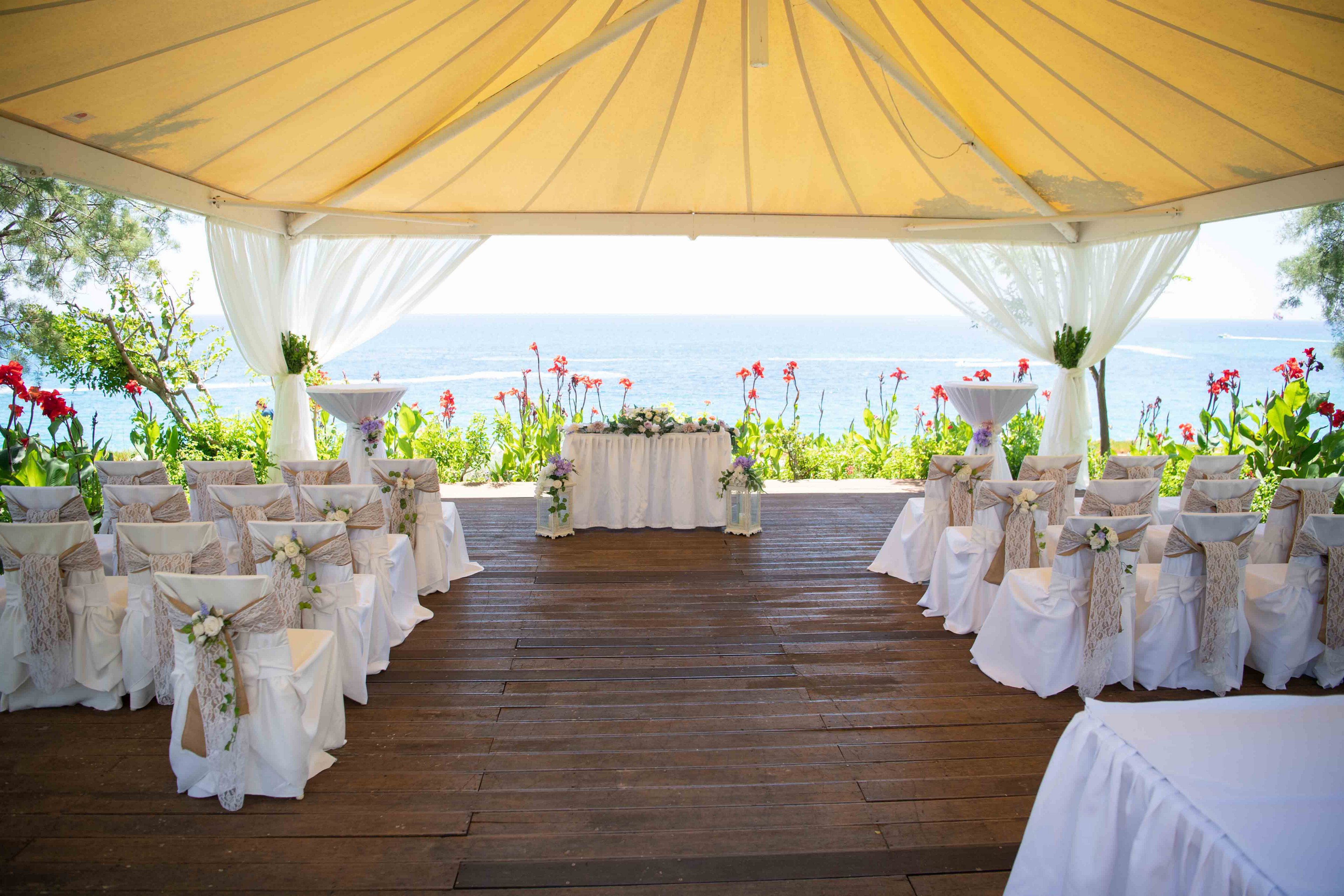 An elegant wedding ceremony area is arranged under a large canopy overlooking the sea. White chairs with decorative sashes are lined in rows on a wooden deck facing a flower-adorned altar table. Sheer curtains frame the open sides of the tent, revealing lush greenery and bright red flowers around the perimeter. The scene suggests a romantic, coastal celebration in a bright, sunny setting.