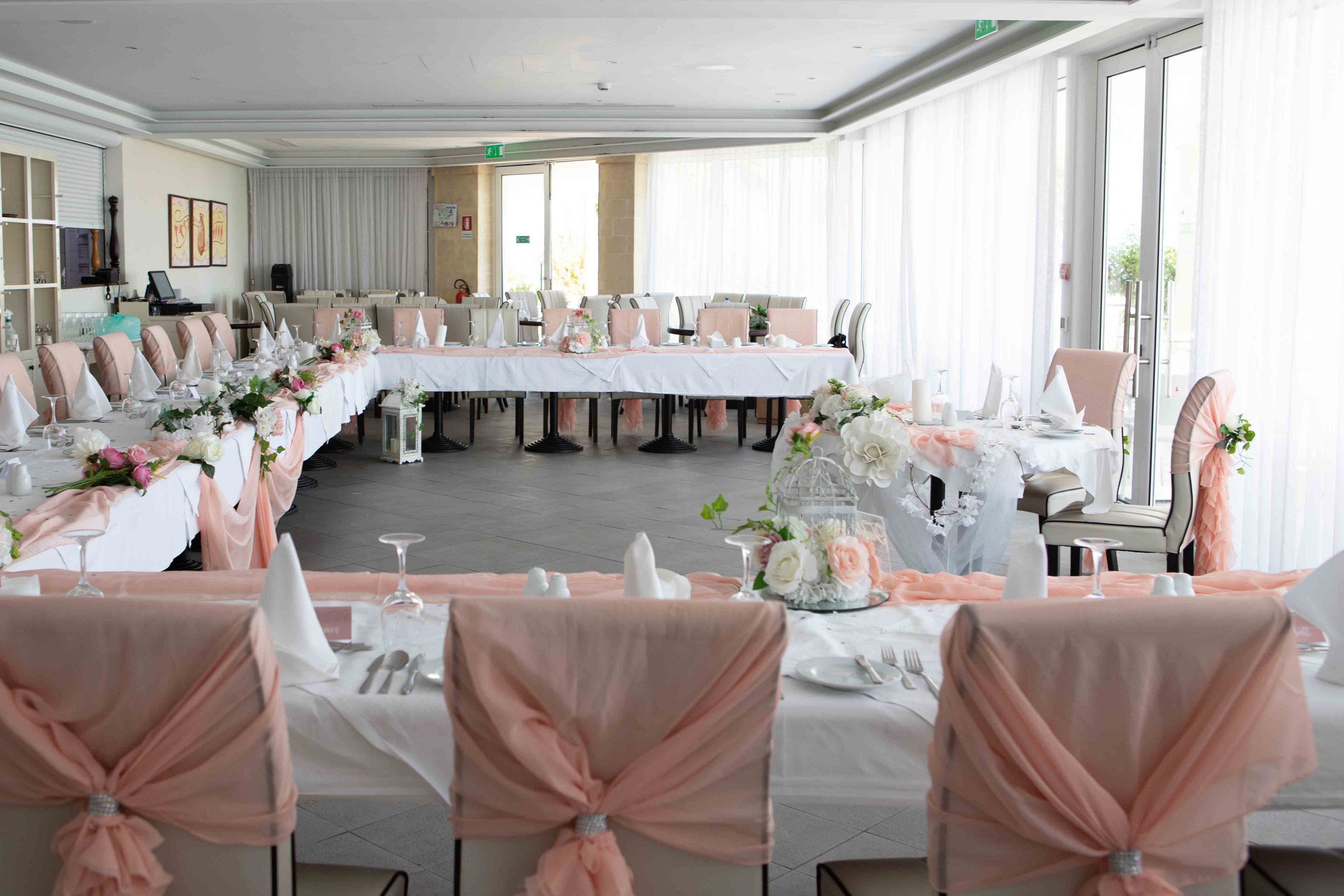 A large banquet hall is arranged with U-shaped tables prepared for a formal event, likely a wedding reception. Chairs are decorated with soft peach fabric bows, and white tablecloths create a bright, airy atmosphere. The tables are set with glassware, folded white napkins, and floral centerpieces featuring white and pink flowers. Sunlight filters through sheer curtains along the windows, enhancing the clean and elegant look of the venue.